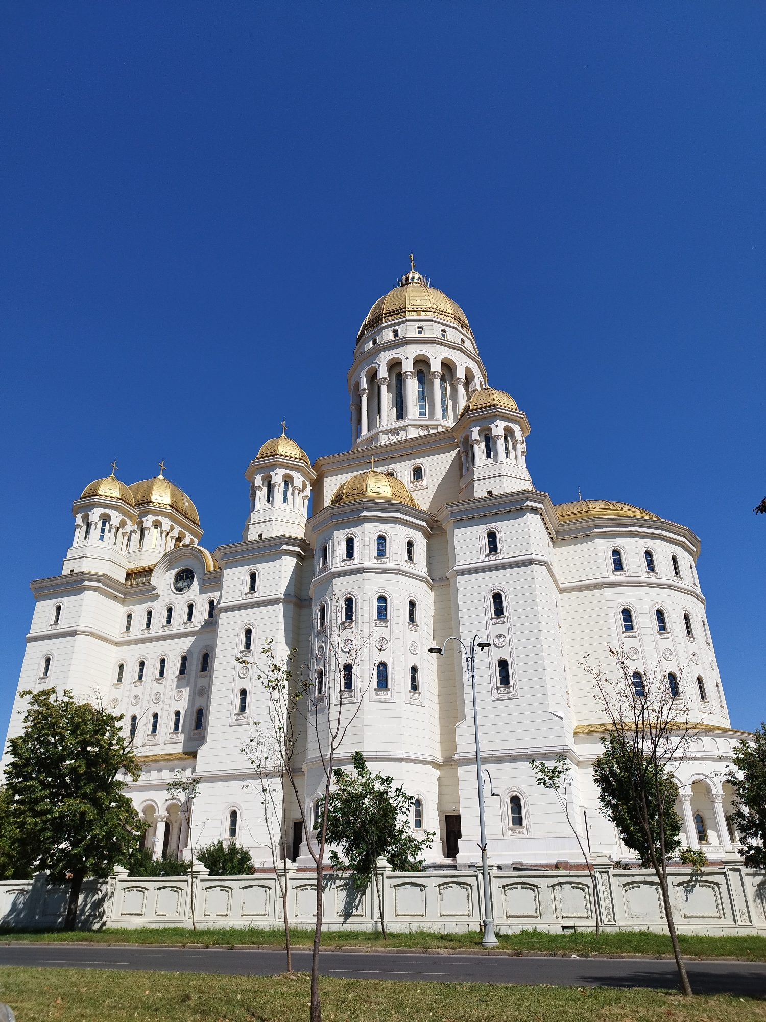 The image shows the exterior of the Romanian Orthodox Cathedral in Bucharest, Romania, on a clear day with a bright blue sky. The cathedral is a large, white building with multiple golden domes and intricate architectural details. The perspective is low-angle, looking up at the cathedral from a street level.
In the foreground, there is a section of a paved road and a grassy area with some trees. A white decorative fence runs along the edge of the cathedral grounds. A lamppost is visible in front of the building. The cathedral itself is the dominant subject, with its imposing facade, numerous windows, and arched doorways. The golden domes catch the sunlight. The surrounding environment appears to be an urban setting, given the road and the presence of trees, but the focus is overwhelmingly on the magnificent architecture of the cathedral. There are no people visible in the image.