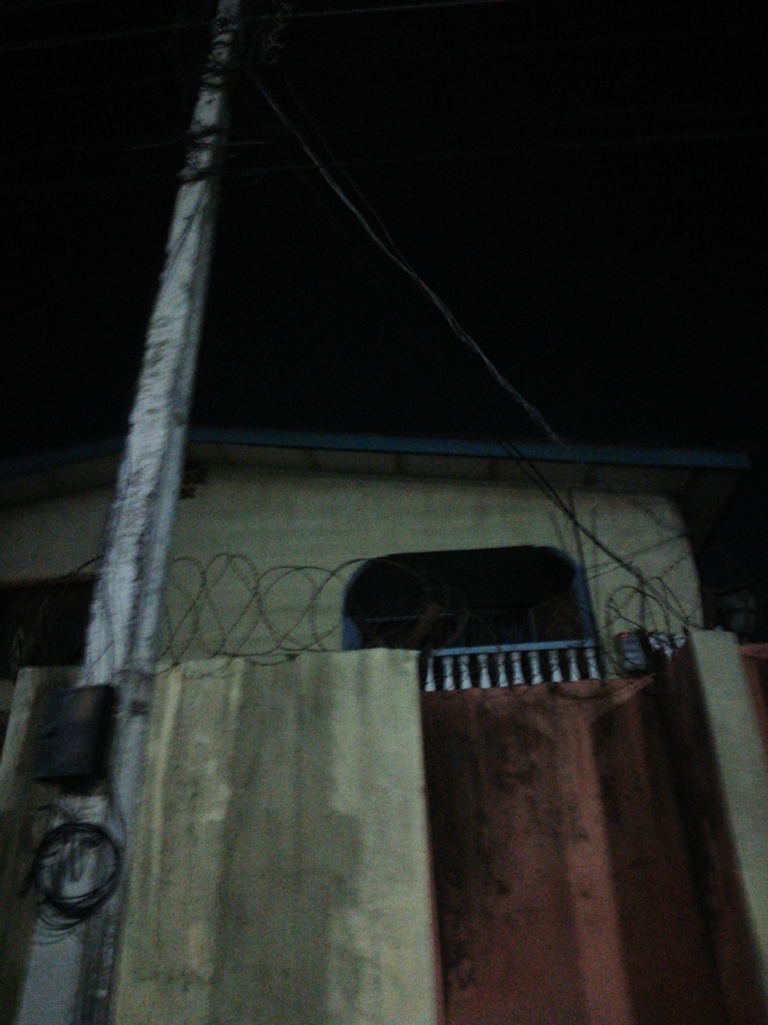At night in Lagos, Nigeria, a grainy, low-angle shot reveals a concrete wall topped with barbed wire. Behind the wall is a tan, two-story building with a blue trim. The building features an arched window or opening with a white railing. A utility pole stands prominently in the foreground, partially obscuring the building. Electrical wires run across the scene. A black electrical box is visible on the wall near the utility pole. The overall setting appears to be an urban residential area, and the barbed wire suggests security concerns.