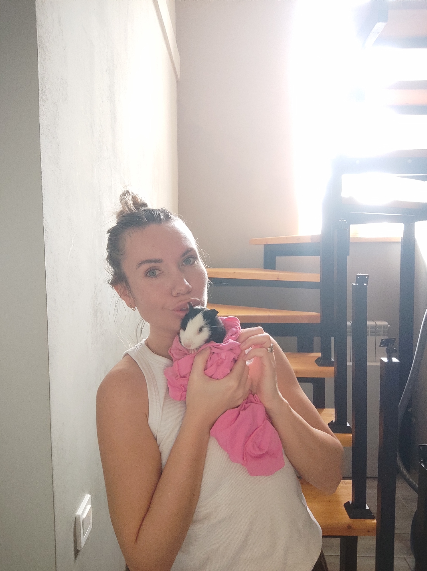 A young woman with light skin, her hair pulled into a loose bun, is holding a guinea pig wrapped in a pink cloth. The woman is wearing a white ribbed tank top. She is indoors, near a staircase with wooden steps and a black metal railing. Natural light streams through a window behind the stairs. A white electrical outlet is visible on the wall to the left. A white radiator is visible behind the staircase. The scene suggests a residential environment in the city of Votkinsk, Russia.