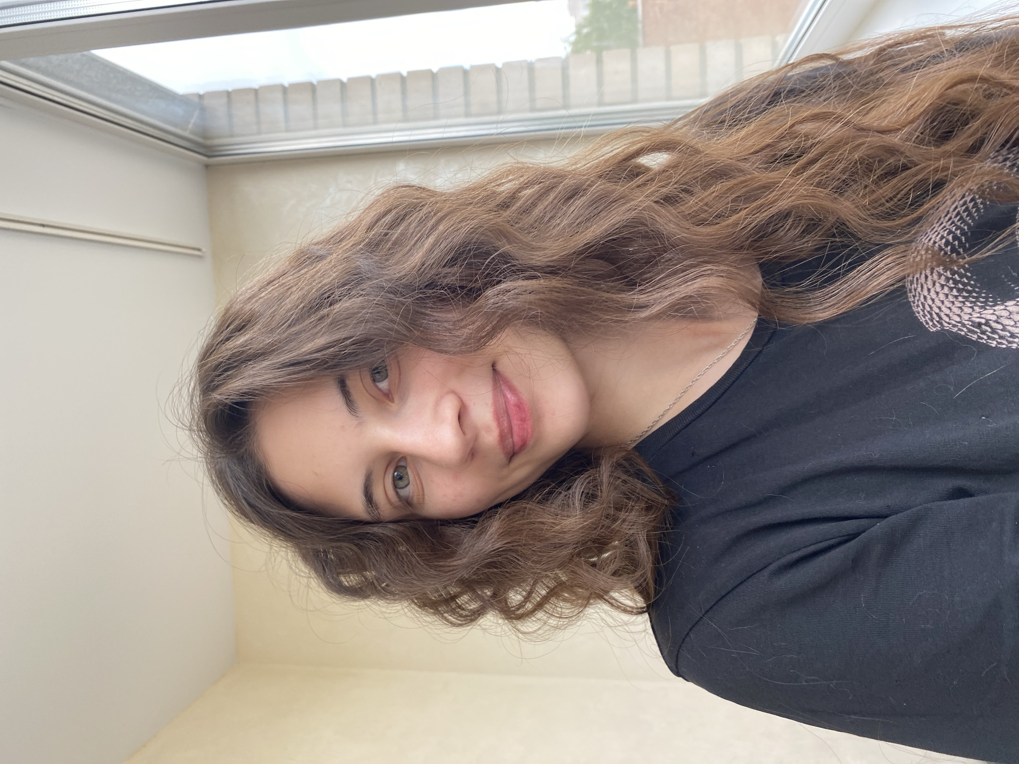 The image depicts a young woman with light brown, wavy hair and fair skin. She is wearing a black t-shirt and a silver necklace. Her eyes are green and she has a slight smile. The background is a room with light-colored walls and a skylight. The image's location in Votkinsk, Russia, is not discernible from the image itself.