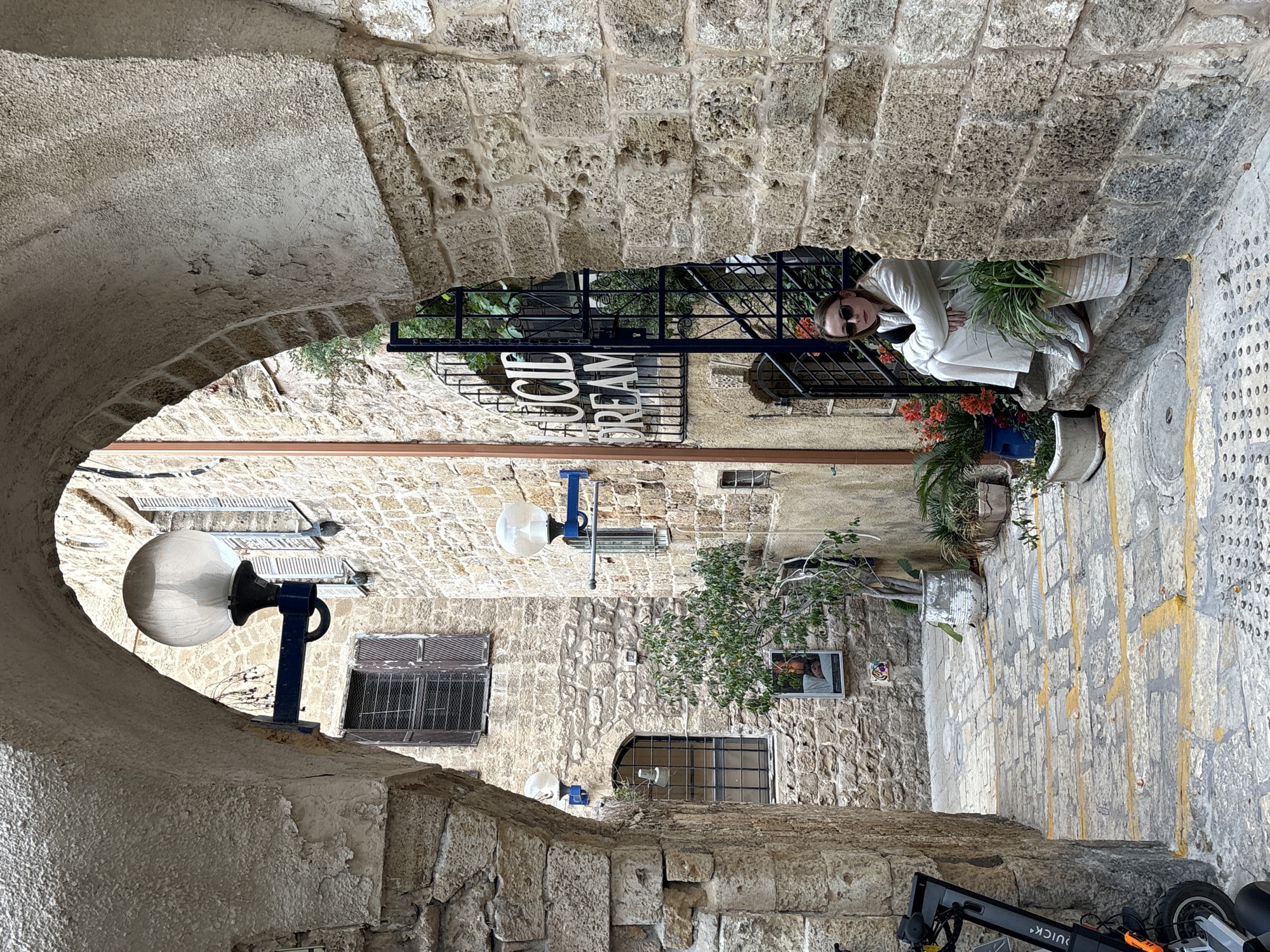 Captured through a stone archway, the scene presents a narrow street in the city of Tel Aviv, Israel. The street is paved with stone and bordered by stone buildings. A woman wearing sunglasses and a white coat is seated near a potted plant adjacent to a black iron railing. A sign reading "Cold Dream" is mounted on the railing. Street lamps are attached to the buildings, and barred windows can be seen. A scooter is parked on the street at the bottom of the frame.