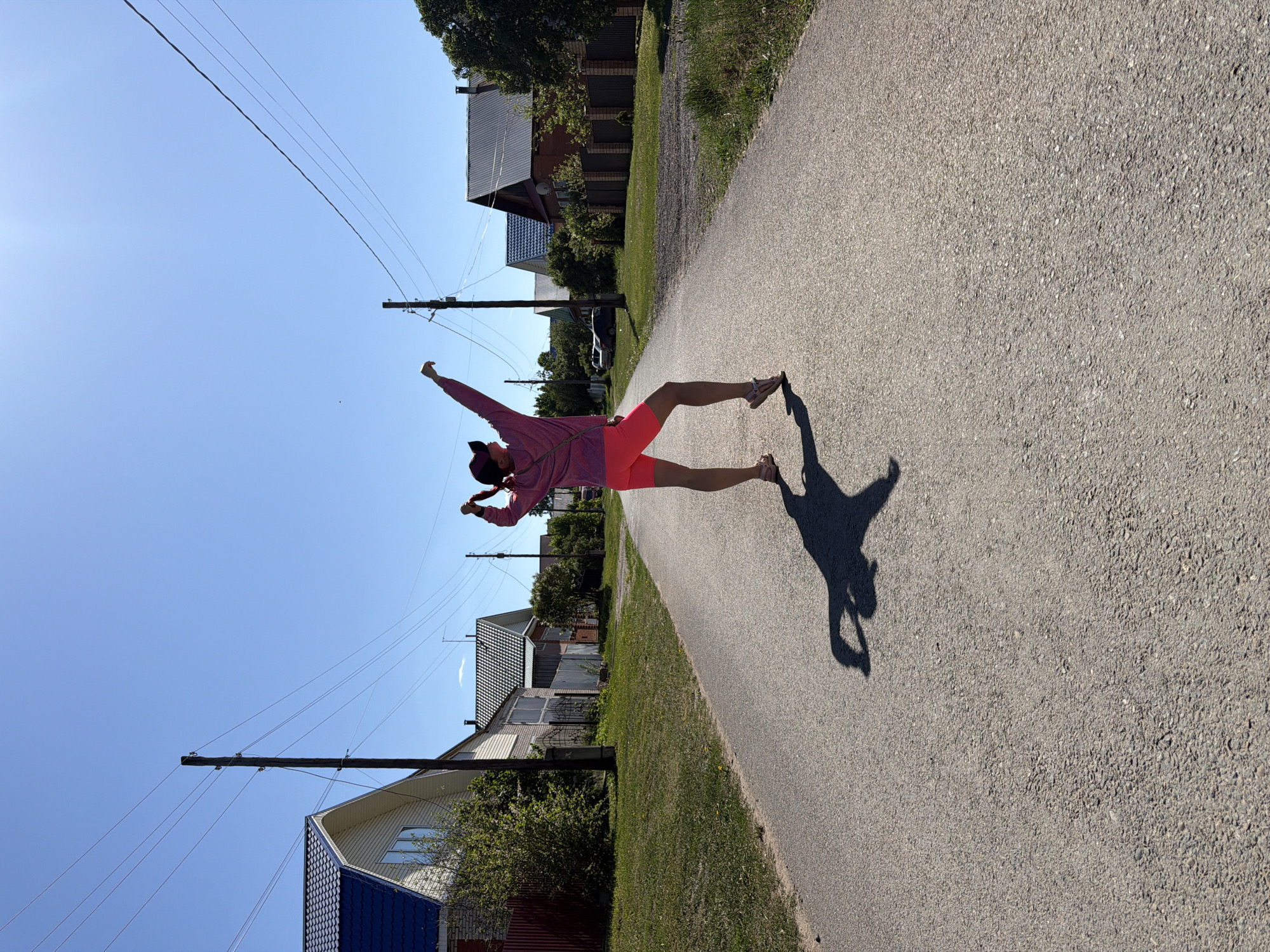 A slightly skewed, medium-distance shot depicts a woman standing in the middle of an asphalt road in what appears to be a residential area. She is wearing a pink jacket and shorts, and a cap, with her arm raised in the air. Her shadow stretches out in front of her on the road. The location appears to be in Chaikovsky, Russia due to the presence of houses with distinctive Russian architecture. Power lines are visible overhead. Houses line the sides of the road, with greenery bordering the asphalt. The sky is clear and blue.