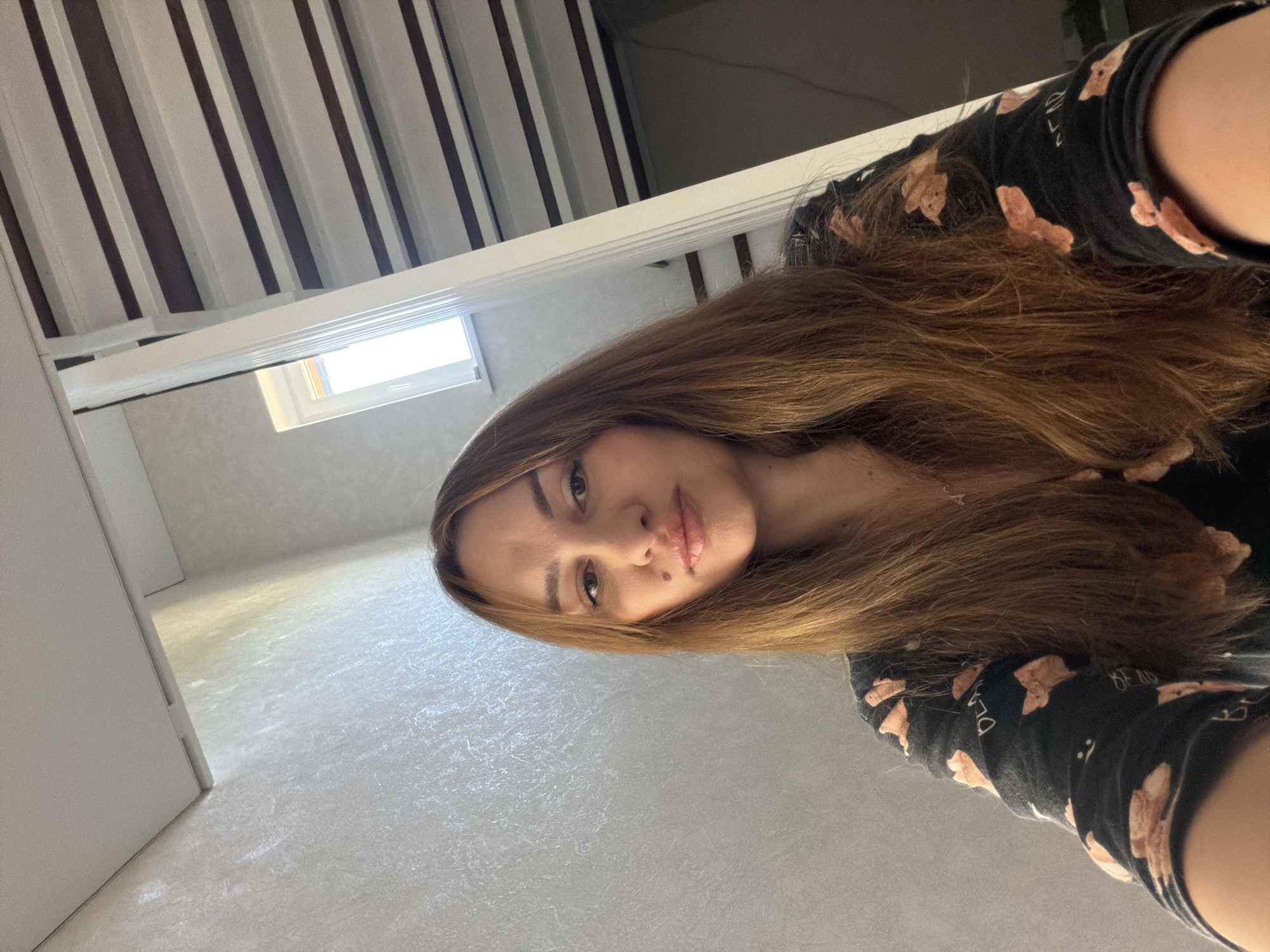 A woman with long brown hair is taking a selfie in an indoor setting, likely a staircase or hallway with a slanted ceiling. She is wearing a black pajama top with a pattern of light brown teddy bears. The walls are textured and light-colored. A small window is visible in the background, letting in natural light. The ceiling above appears to be made of wooden beams and white planks. There is no information in the image to suggest this location is in Malinovka, Russia.