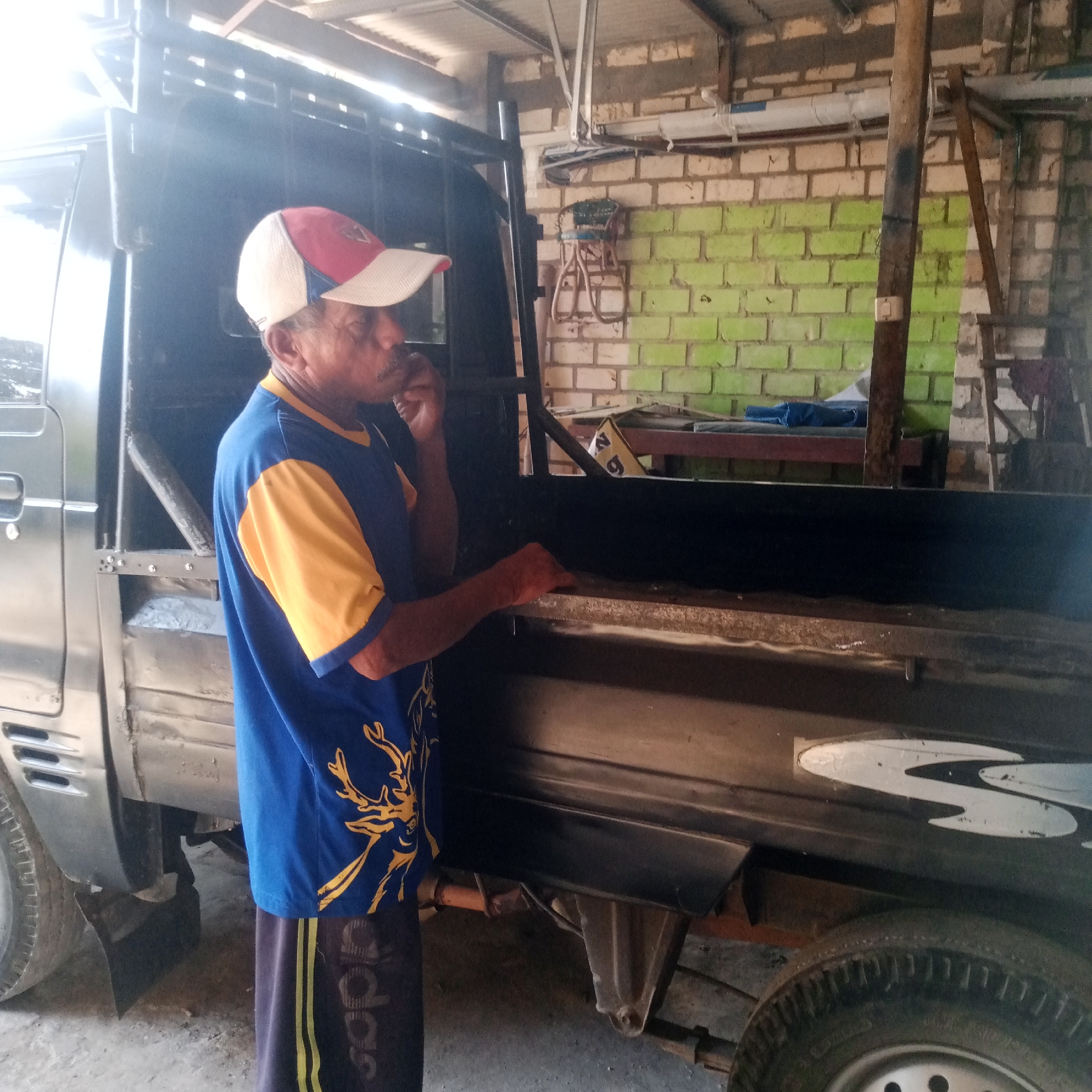 An older man wearing a red, white, and blue baseball cap and a blue t-shirt with a yellow collar and yellow sleeves, emblazoned with a gold deer graphic, stands next to the open bed of a dark gray pickup truck. He has a mustache and is bringing his right hand to his mouth. The truck bed appears to be made of metal and has some debris inside. The background shows a brick wall painted in sections of white and lime green, a wooden post, and some shelving with items on it. The ground is dusty. The scene appears to be a workshop or garage. The image was taken in Semanding, Indonesia.