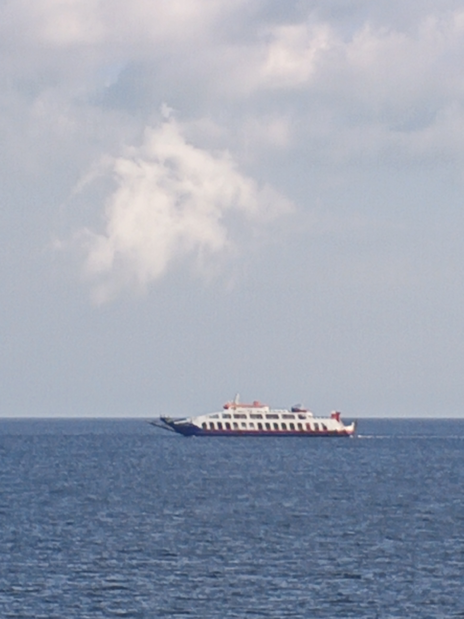A ferry is sailing on the sea under a cloudy sky. The ferry is white with red trim and appears to be a passenger or vehicle ferry. The water is a deep blue and appears choppy. The sky is a pale blue with scattered white clouds. The image was taken from a distance, and the ferry is the main subject. The location is unknown, Indonesia.