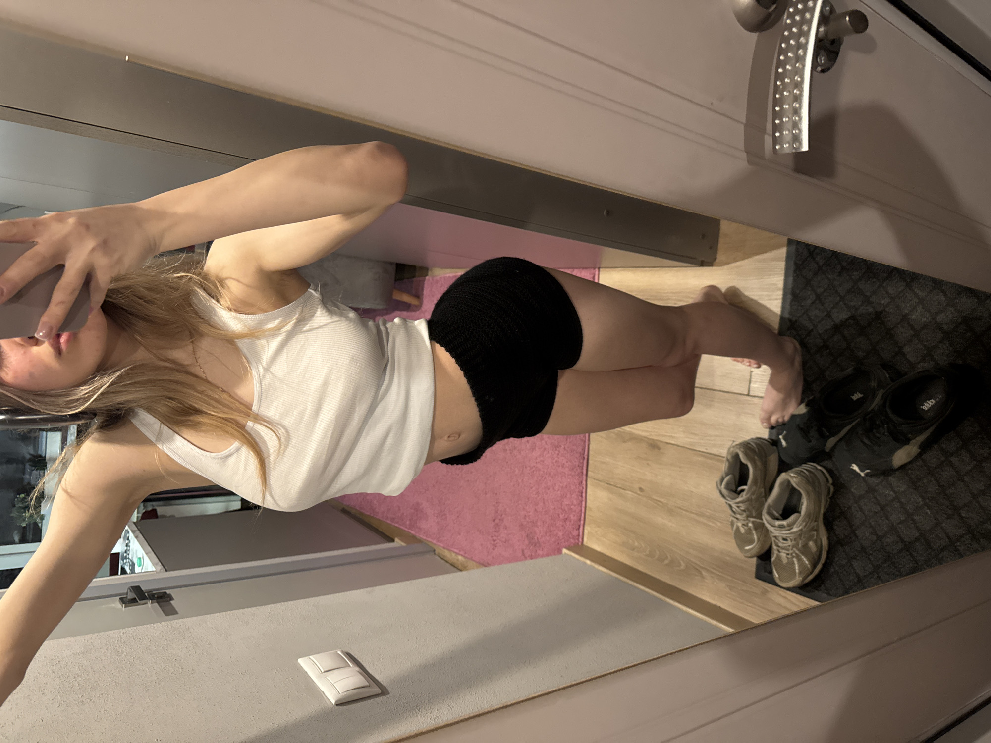 A young woman with blonde hair is taking a selfie in what appears to be an apartment. She is wearing a white tank top and black shorts. The room has light grey walls and a pink rug. There are several pairs of shoes near the doorway. The location is in Khimki, Russia.