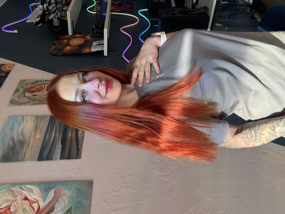 The image depicts a female subject in an indoor residential setting. The location, Rostov-on-Don, Russia, cannot be confirmed from the visual evidence provided.
The subject is a woman with long, bright reddish-orange hair and blue eyes, smiling faintly and looking directly at the camera. She has a silver nose ring on her right nostril. Her left hand is placed on her upper chest, revealing a faint tattoo on her thumb. Her right forearm is heavily tattooed with intricate patterns, including what appears to be a moth or butterfly and floral designs. She is wearing a light grey short-sleeved t-shirt, a silver chain necklace with a small cross pendant, and a white smartwatch on her left wrist. Strong, warm light illuminates her from the left, highlighting her hair and face, and creating a noticeable sheen on her shirt.
The background consists of two distinct wall sections. To the left, a textured, light beige wall displays three vertically aligned framed artworks: a large pink floral painting, a seascape depicting a sunset over waves, and an abstract piece with a reddish-brown geometric form. To the right, a darker, possibly navy blue, wall section is angled, suggesting a corner. This section features two light-colored floating shelves. The top shelf holds a detailed model, possibly a motorcycle. The bottom shelf displays a black camera, a blue model car, and other small items. A wavy, multi-colored LED strip light runs vertically down this darker wall. The lower right portion of the image reveals a partial view of a desk area with a black computer speaker and a monitor.