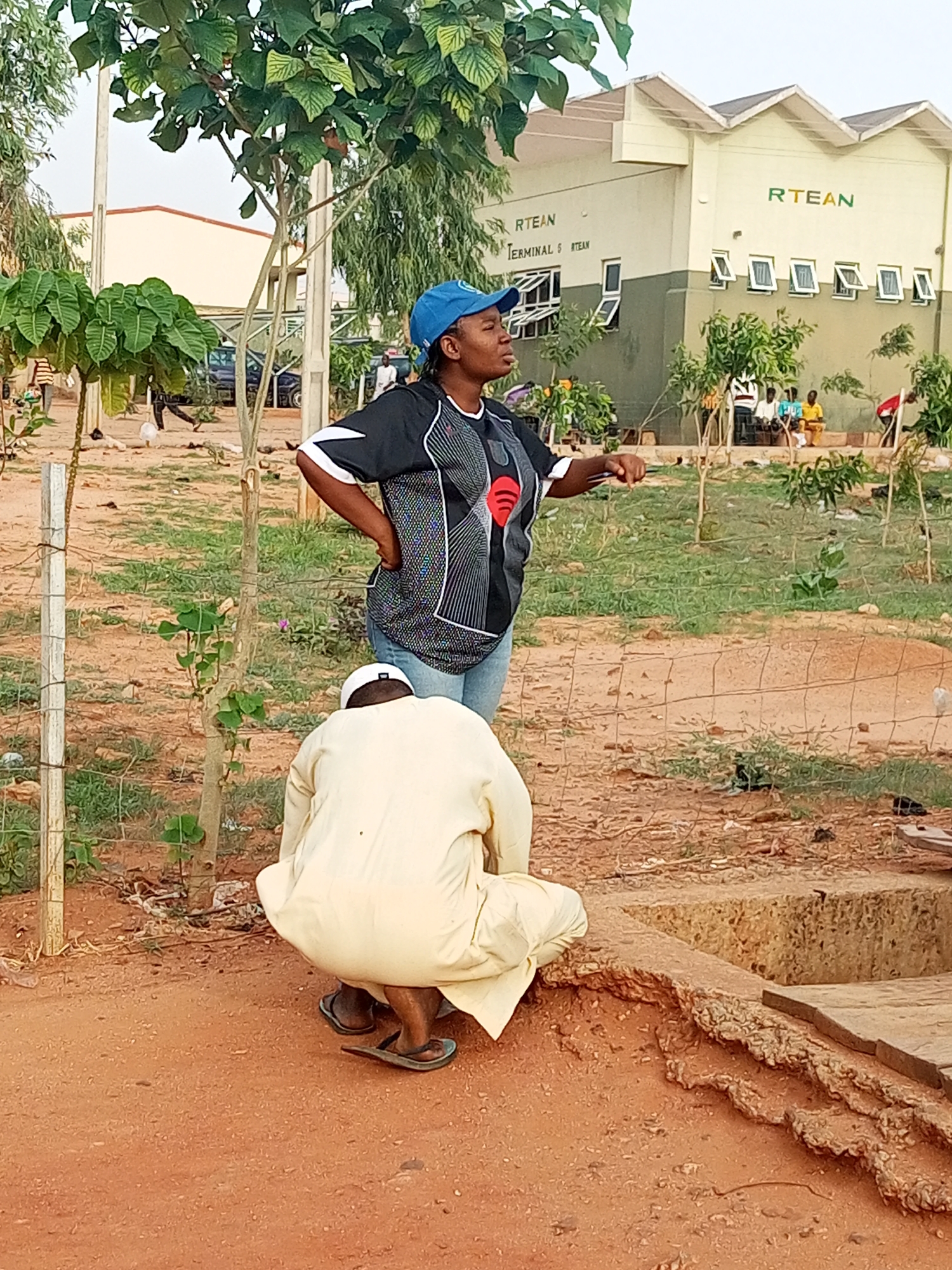 In an outdoor setting in Gombe, Nigeria, a woman wearing a blue cap, a black and white patterned shirt with a red Spotify logo, and blue jeans stands with her hand on her hip, gesturing with her other hand. A man wearing a white outfit and a white cap is squatting in front of her, near a low concrete structure. The ground is reddish-brown dirt. Behind them is a grassy area with sparse vegetation and a wire fence. In the background, a building with "RTEAN TERMINAL 5 RTEAN" written on it can be seen. Other people are visible in the background.