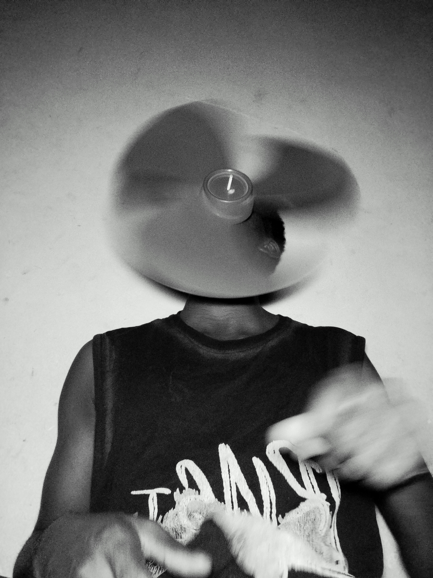 In a black and white image, a person is positioned in the center, facing forward. They are wearing a dark, sleeveless t-shirt with white lettering on the front. Their head is obscured by a round object that is in motion, creating a blurred effect. A small, circular object with the numeral "1" is visible on the spinning object. The person's arms are also blurred, suggesting movement. The background is a plain, textured wall. The lighting appears to be from above and slightly to the left, casting a shadow of the spinning object on the wall. The overall impression is one of dynamic movement and abstraction.