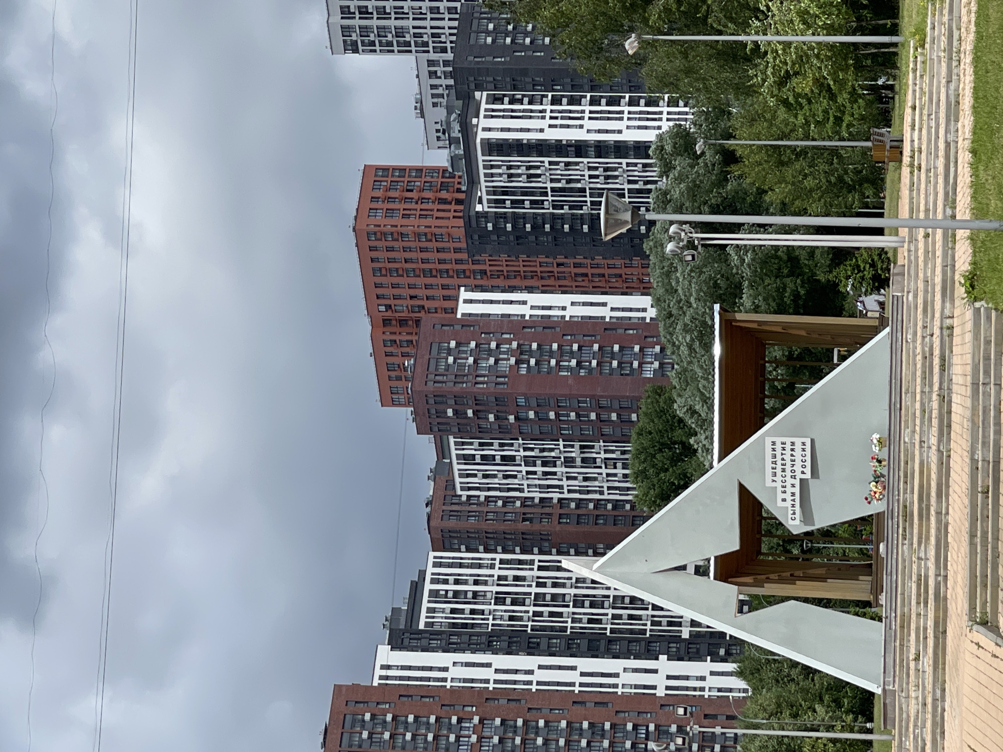 The image captures a view of the Eternal Flame monument in Moscow, Russia, situated in front of a backdrop of modern residential high-rise buildings. The monument, an abstract triangular structure, features a prominent star at its apex. White text on a darker background within the monument reads, "УШЕДШИМ В БЕССМЕРТИЕ СЫНАМ И ДОЧЕРЯМ РОССИИ" (To those who departed into immortality, sons and daughters of Russia). Several floral arrangements are placed at the base of the monument. The surrounding environment includes lush green trees and multiple lampposts. The sky is overcast with heavy clouds, suggesting a gloomy or rainy day. The high-rise buildings are a mix of brick and modern facades, with varying colors of red, white, and dark gray. The scene appears to be a public space, possibly a park or a memorial ground, within a city.