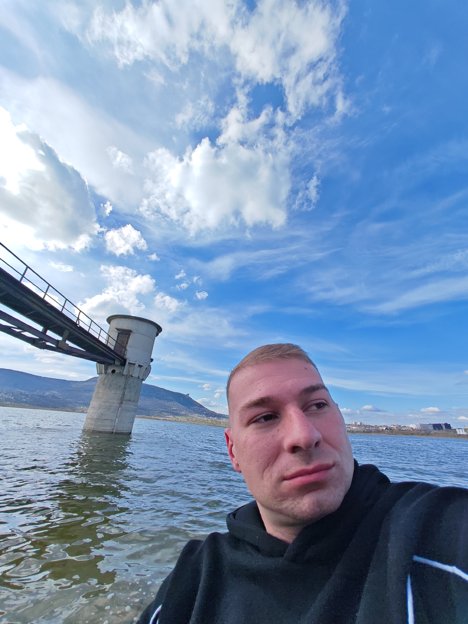 A man stands in water, possibly a lake or river, with a gray structure behind him that appears to be a pumping station or water intake tower. The structure is connected to land by a metal walkway or bridge. The man is wearing a black hoodie and appears to be taking a selfie. The sky is blue with scattered clouds. The water is choppy, and there are hills or mountains in the background. The photograph was taken at the Yazovir Ticha (Ticha Dam), in Shumen, Bulgaria. The location is near a bridge.