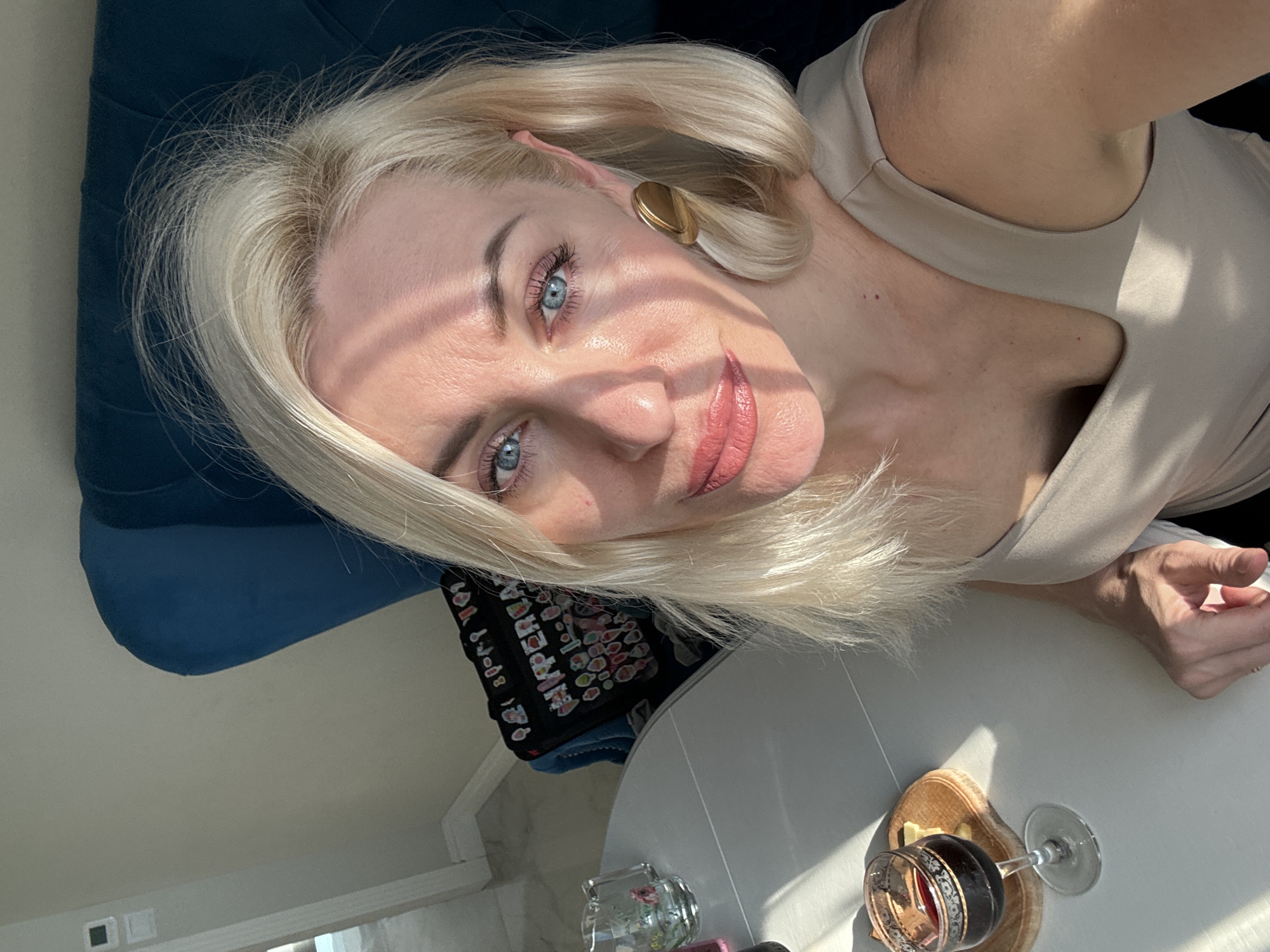 A fair-skinned woman with blonde hair and blue eyes is indoors, sitting at a white table. She is wearing a light beige top and gold disc earrings. A glass of red wine is on the table near her hand. A blue upholstered chair or cushion is visible in the background. The location is Unknown, Unknown.