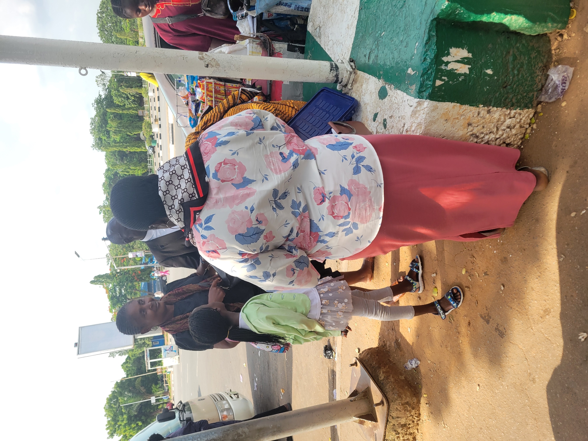This outdoor image, set in Abuja, Nigeria, captures a vibrant street scene on a sunny day. The foreground is dominated by a woman viewed from behind, wearing a white blouse adorned with pink and blue floral patterns, a long red skirt, and a black headscarf featuring a white geometric print. She stands near a roadside stall displaying various packaged goods, including snacks and beverages, alongside a yellow woven item and a blue plastic tray.
Beside her stands a young girl, dressed in a light green jacket over a white top, a grey tulle skirt, and light grey leggings, complete with blue and white sandals. Another woman, wearing a dark top and a patterned scarf, is visible in the mid-ground, partially facing the camera with a gentle smile. Further back, several other individuals are present, including one person in traditional red attire near a white van on the street.
The scene features a prominent white pole on the left and a distinctive green and white painted concrete curb along the roadside. Lush green trees line the background, and the ground is a mix of dirt and sand with scattered small debris. The bright, clear daylight and absence of shadows directly under feet suggest a mid-day or early afternoon setting. Visible text on a blue package near the stall reads "DEN PEN." The overall impression is one of everyday street activity in an urban environment.