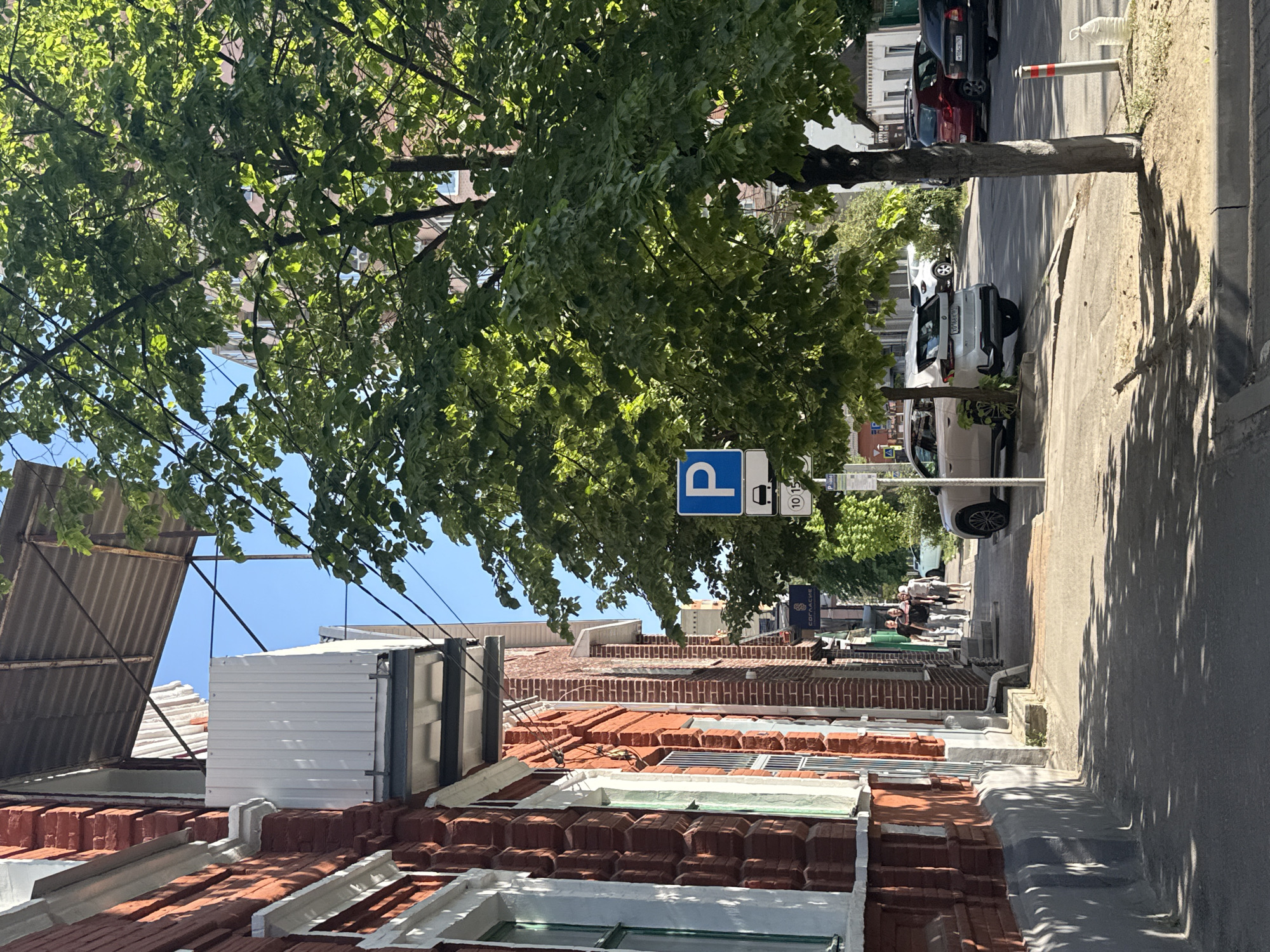 The image shows a street in Rostov-on-Don, Russia. The row of buildings along the street features red brick construction with white trim around the windows and along the roof edges. On the roofs, there are white and gray metal structures. A parking sign is visible on the right-hand side of the road. Cars are parked along the street, including a white SUV. The street is lined with trees. A group of people is walking on the street in the distance.