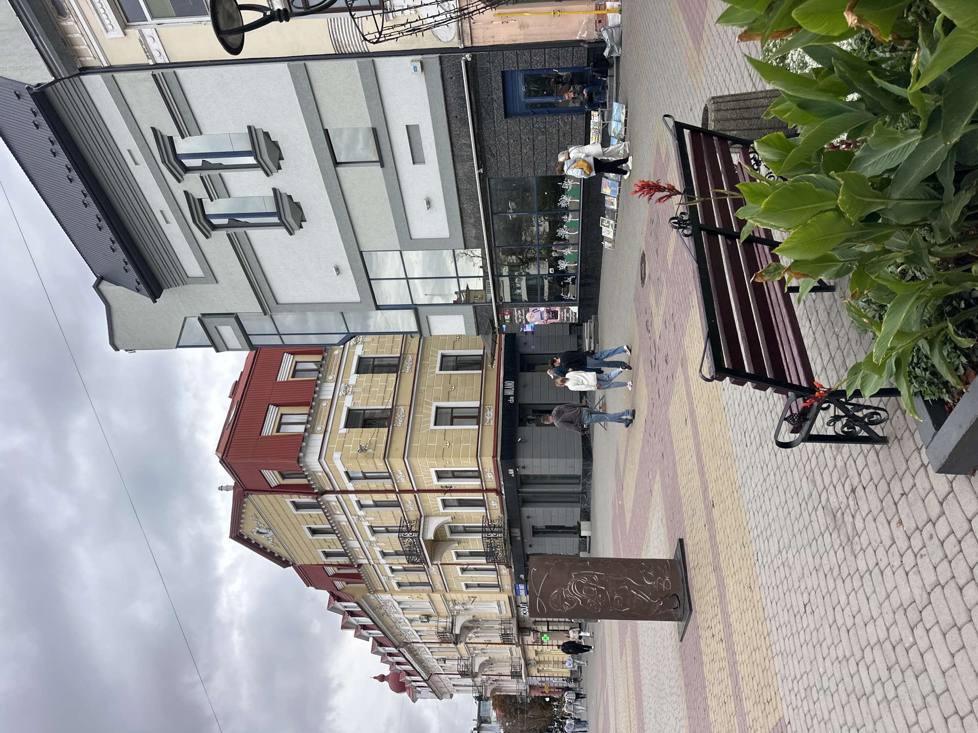 The image depicts a street scene in Ternopil, Ukraine, featuring a pedestrianized area with several buildings. On the left, a row of traditional buildings with yellow facades and red roofs are visible, characterized by balconies and ornate architectural details. To the right, a more modern building with a grey facade and large glass windows stands out.
In the foreground, a patterned paved walkway is observed, with a dark metal bench in the lower right quadrant. A metal sculpture or monument is positioned in the middle ground. Several people are walking along the street, some appearing to be shoppers or tourists. There is an outdoor display of artwork in front of one of the ground-floor establishments. The sky is overcast, suggesting a cloudy day. The overall atmosphere appears to be that of a bustling urban center.