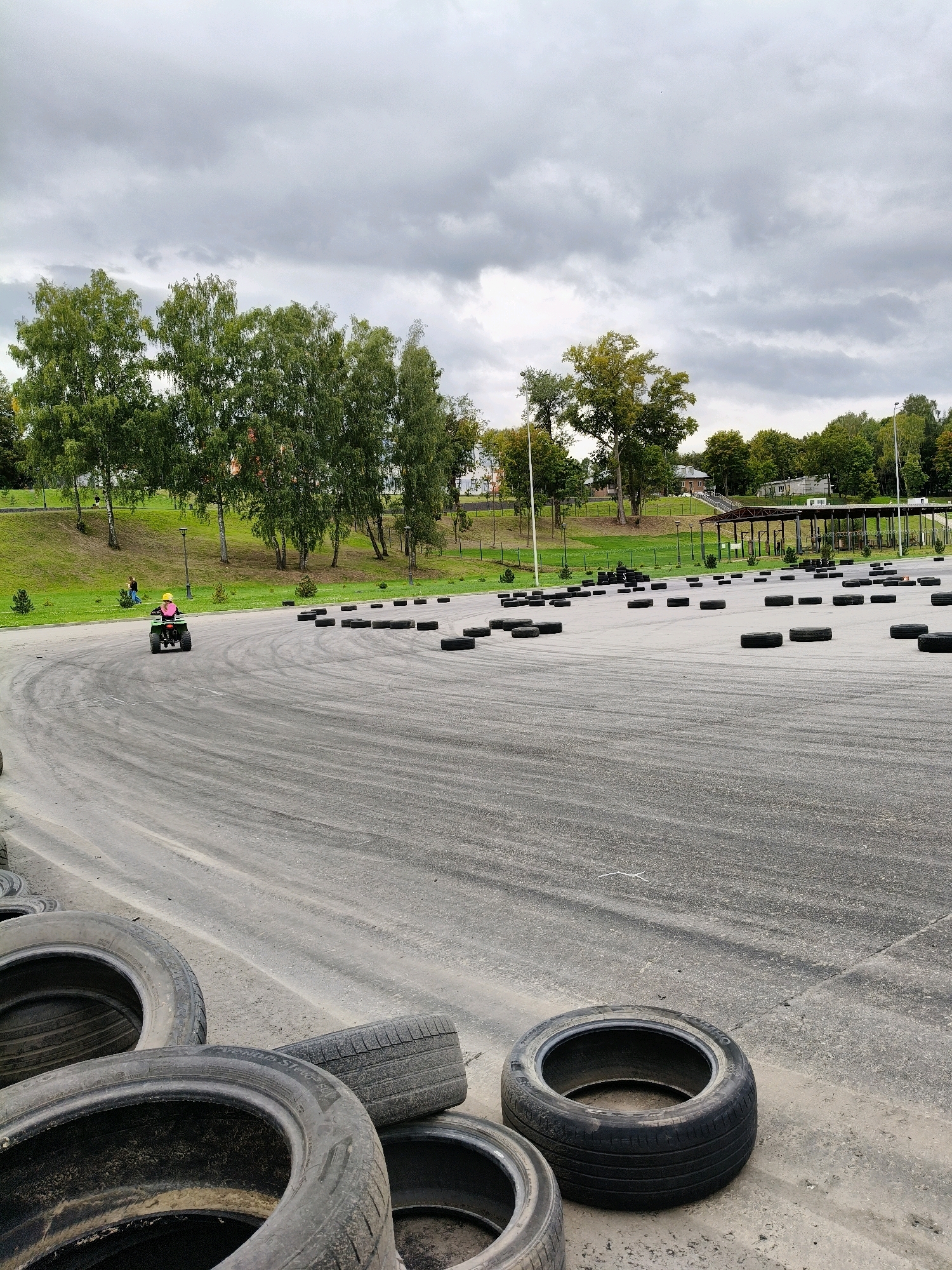 A child is riding an ATV on a track in an outdoor recreation area. The track is made of asphalt and is bordered by grass and trees. Several tires are placed around the track, possibly as obstacles or markers. The sky is overcast, and the overall lighting suggests it is daytime. The child appears to be wearing a helmet. In the background, there are trees and some buildings. The scene seems to be a karting or ATV track.
