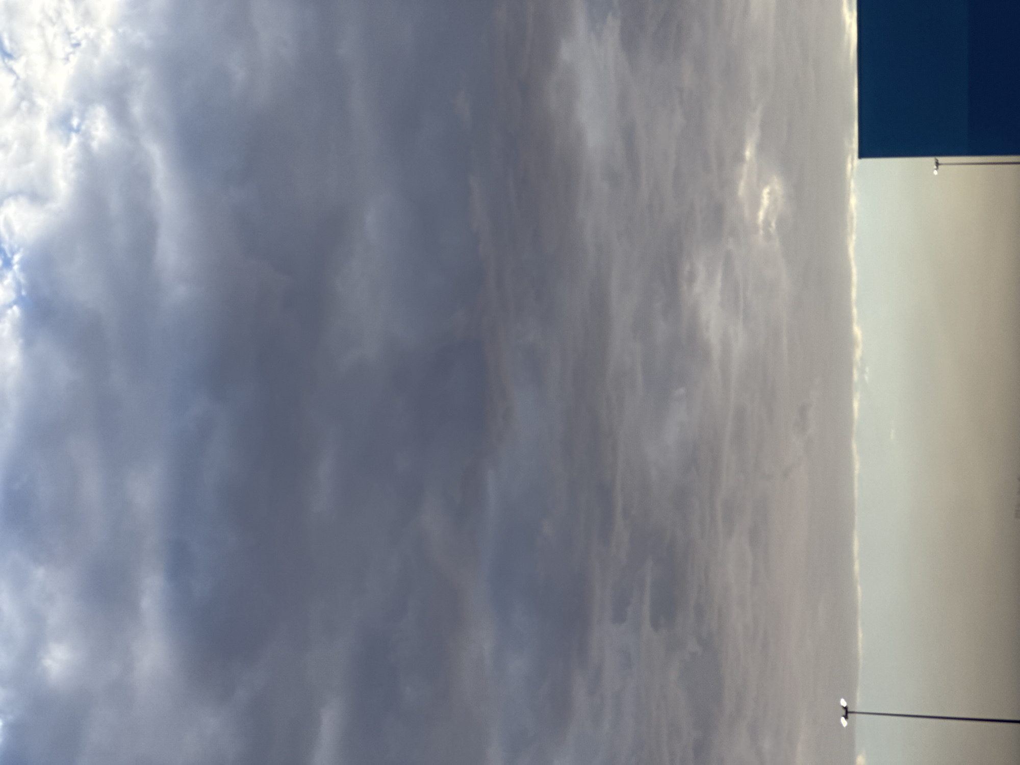 The image shows a cloudy sky over El Paso, Texas. In the lower portion of the image, a dark blue rectangular structure is visible on the right side. A streetlamp with two lights is positioned on the left side of the frame. The sky is filled with various shades of gray clouds, with hints of lighter sky peeking through in some areas. The overall atmosphere suggests an overcast or potentially stormy day.