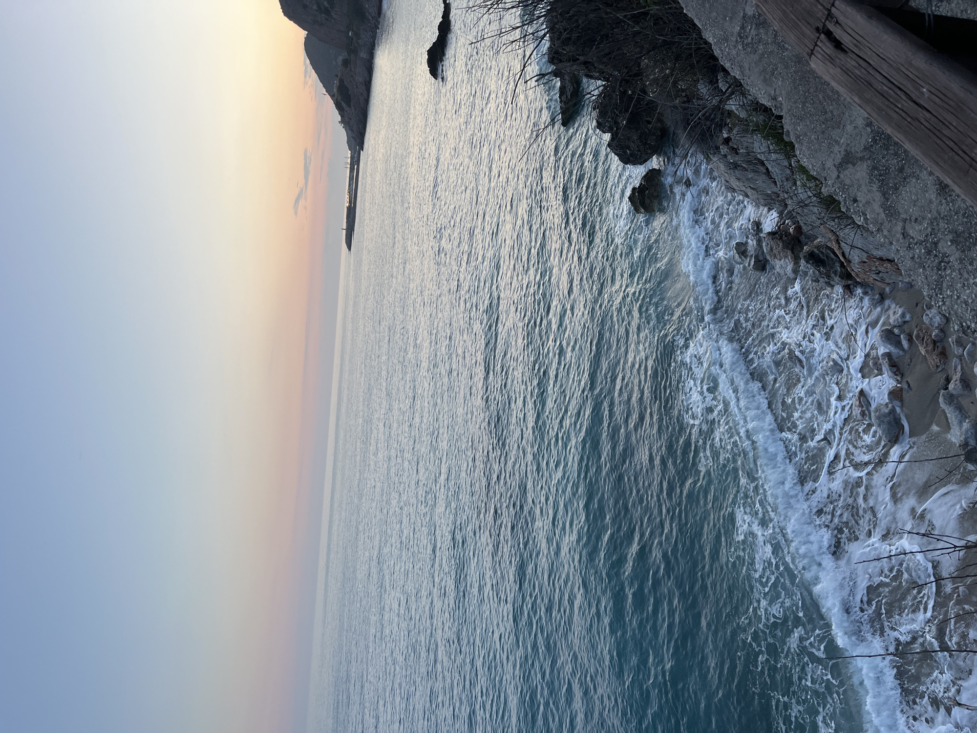 This is a long, landscape-oriented shot of the Mediterranean Sea at sunset from a high vantage point in Alanya, Turkey. The sky transitions from blue to orange to pink. The calm water reflects the colors of the sunset. Along the shoreline, waves crash into a small beach. The edge of a rocky cliff with vegetation is visible in the foreground. In the distance, a pier extends into the water, with a lighthouse at the end.