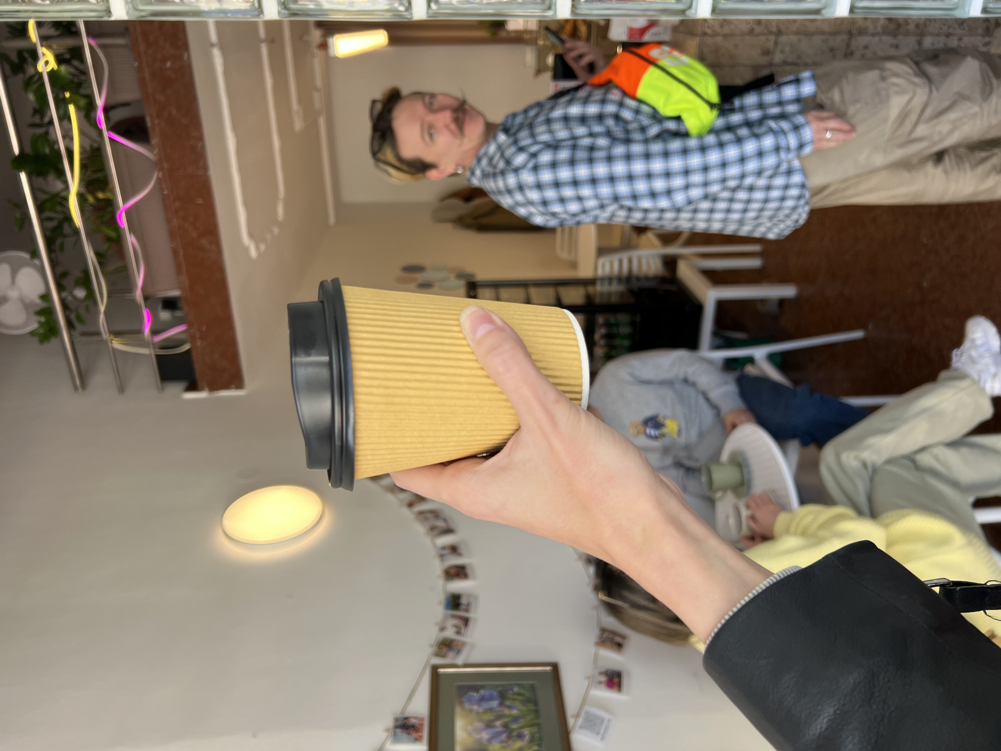 A horizontally oriented image shows a person holding a brown paper coffee cup with a black lid. The person is wearing a black leather jacket and a striped gray-and-white shirt is visible at the wrist.
In the background, another person stands in a blue-and-white plaid shirt and khaki pants. A green and orange vest is visible behind them. A blurred group of people sit at a table. The scene appears to be indoors, possibly a cafe or coffee shop. A wall with framed pictures and string lights is visible. Based on the photo context, the location is likely in Bar, Montenegro.
