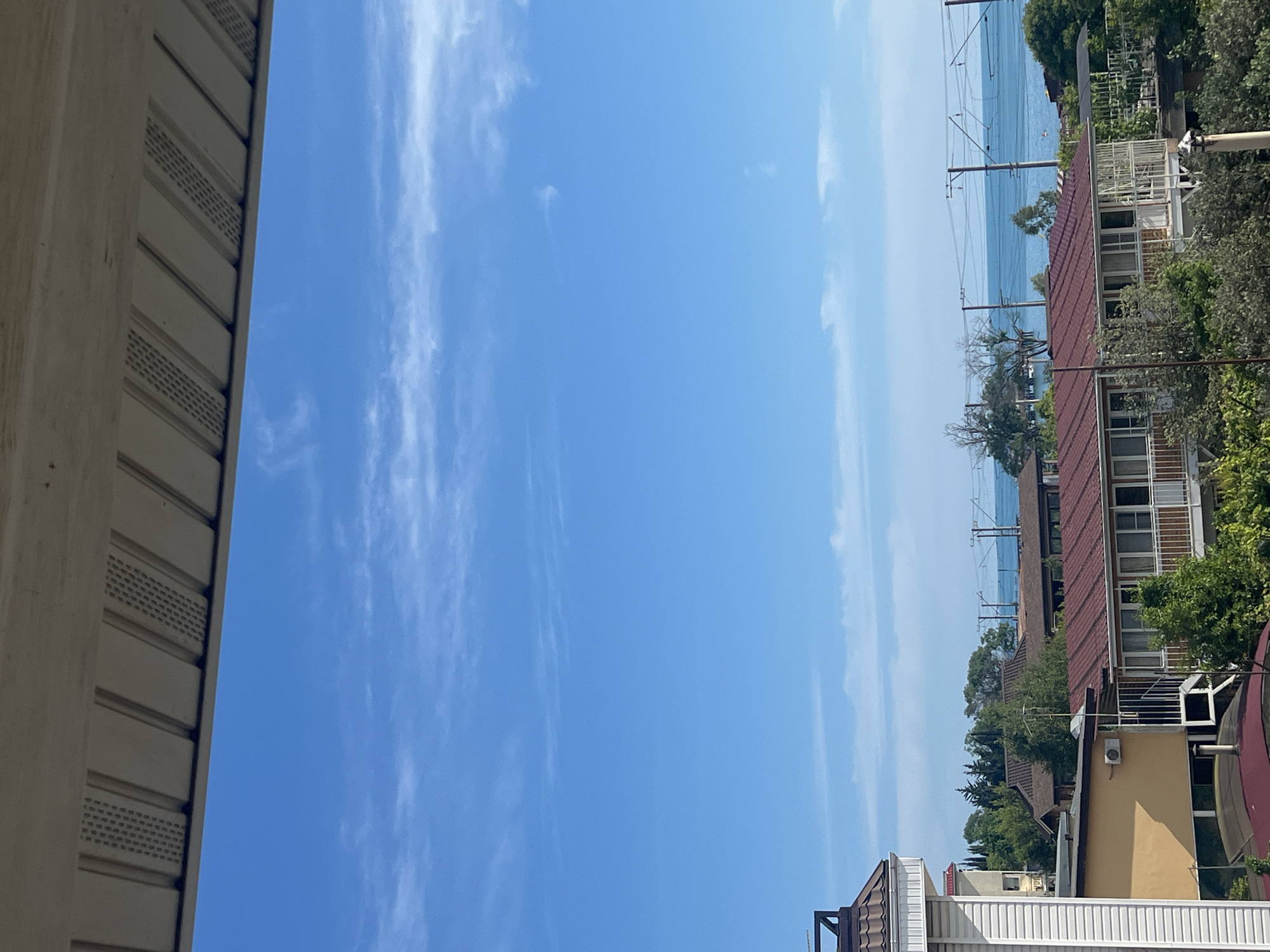The image depicts a view from a balcony overlooking a coastal town, likely in Sochi, Russia, given the architecture and vegetation. In the foreground, a white, weathered soffit with horizontal slats and ventilation cutouts forms the top edge of the frame. Below this, a clear blue sky with sparse, wispy white clouds stretches across the upper half of the image. The middle ground features a cluster of buildings with red tiled roofs, some appearing to be houses or small hotels. One building on the left has a light yellow facade and an outdoor air conditioning unit. Adjacent to it, a multi-story building with white siding and a dark-tiled roof is visible. Lush green trees and foliage surround the buildings. In the background, a calm blue sea meets the horizon, with power lines and poles running horizontally across the scene above the water. There are no people or visible actions taking place.