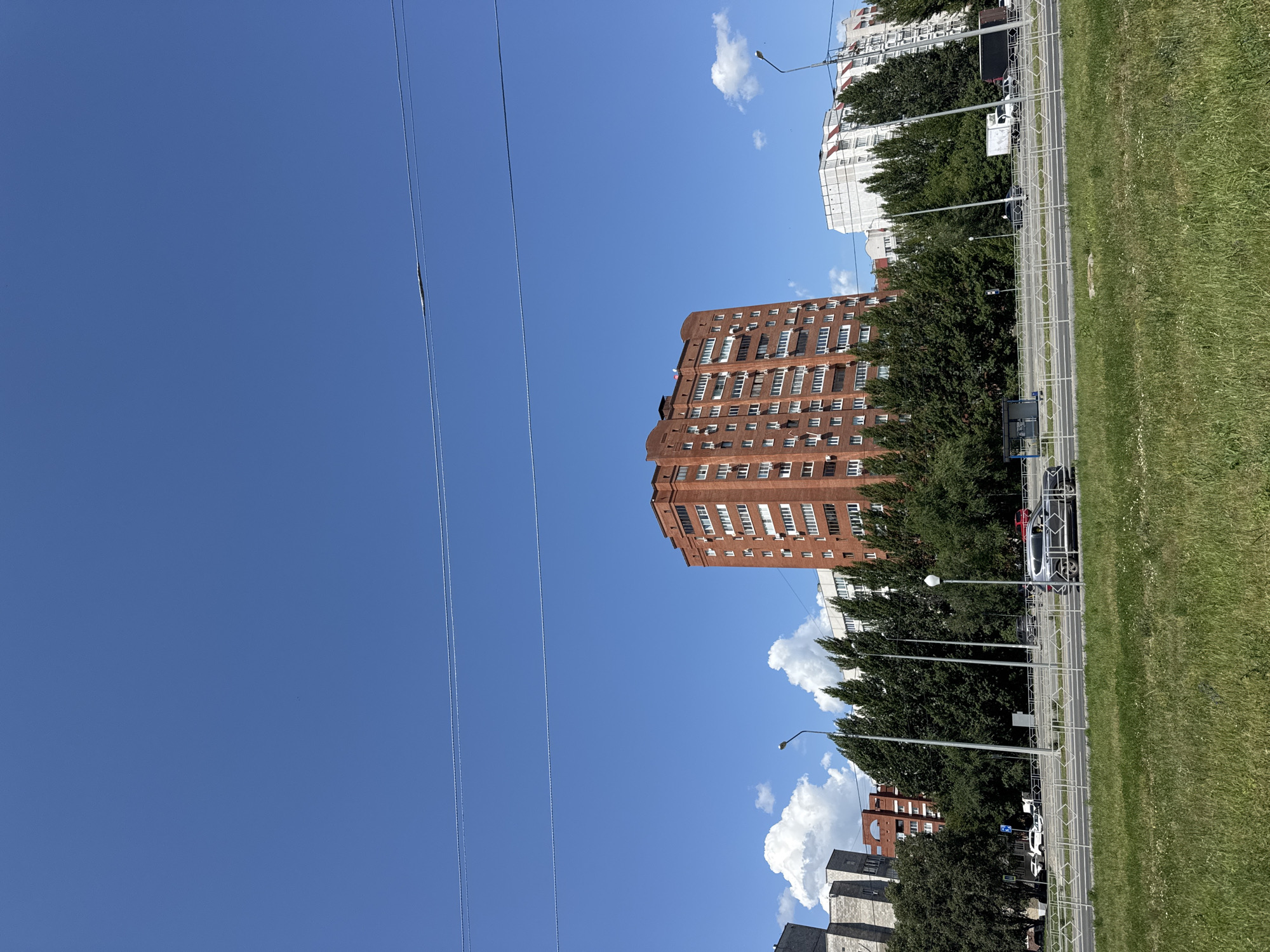 In Tolyatti, Russia, a street scene unfolds under a clear blue sky. A multi-story brick apartment building with numerous windows dominates the central view, flanked by other residential buildings and a line of tall, dark green trees. In the foreground, a strip of green grass with sparse wildflowers slopes gently towards a paved road. Several cars, including a silver SUV and a white truck, are visible on the road, separated from the grassy area by a white metal fence. Overhead, two thin power lines are stretched across the sky.
