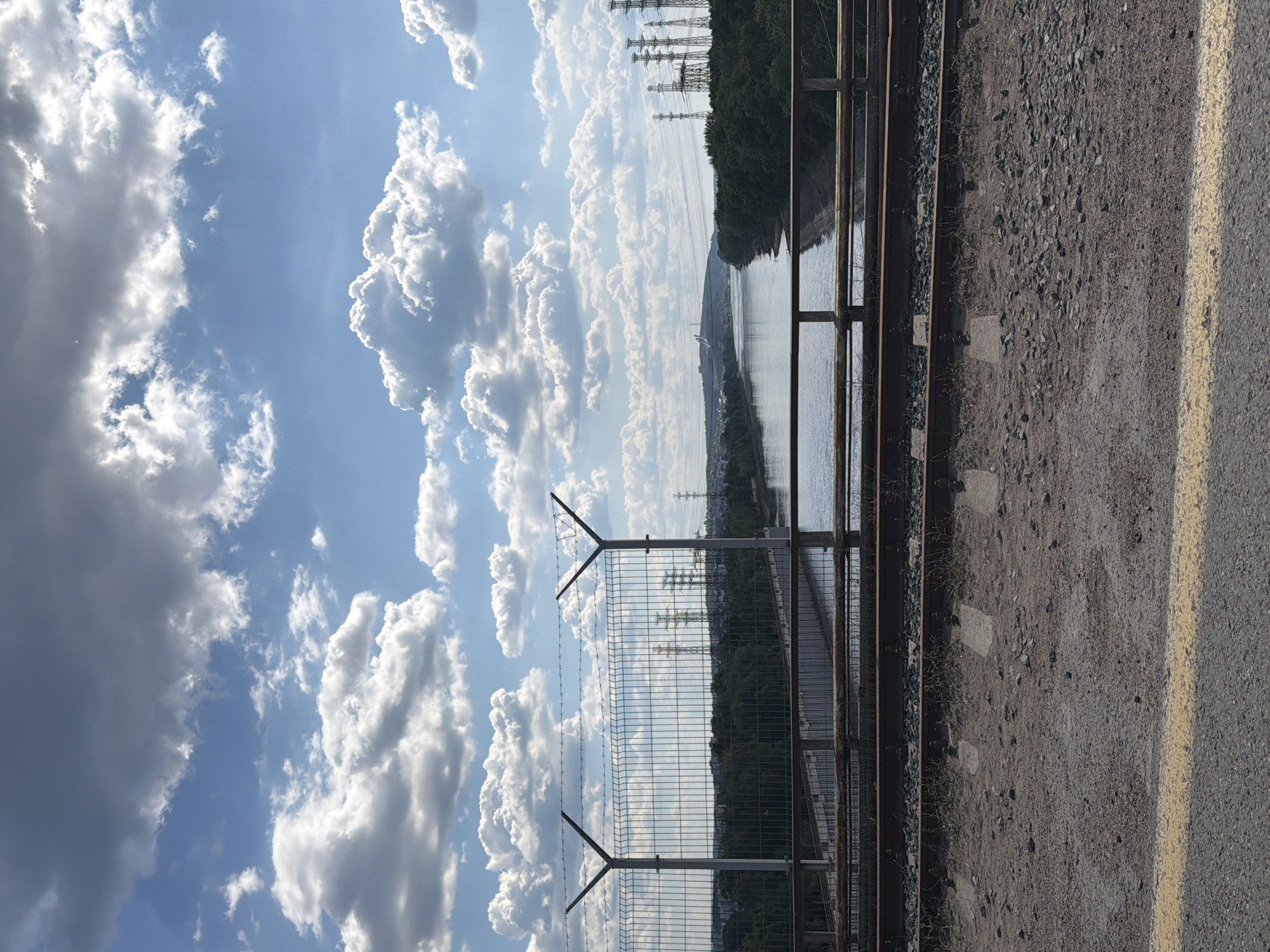The image depicts a wide river flowing through a landscape with a bridge in the foreground and a vast sky filled with fluffy white and grey clouds. The bridge appears to be a railway bridge, with tracks and ballast visible. A fence with barbed wire tops it, suggesting a security measure. Beyond the bridge, the riverbanks are lined with dense green trees. In the distance, several tall electricity pylons stand out against the horizon, indicating industrial activity. The overall scene is one of natural beauty juxtaposed with industrial infrastructure, under a dynamic sky. The location is identified as the Kama River in Chaikovsky, Russia, a city known for its hydroelectric power station. There are no people visible in the image.