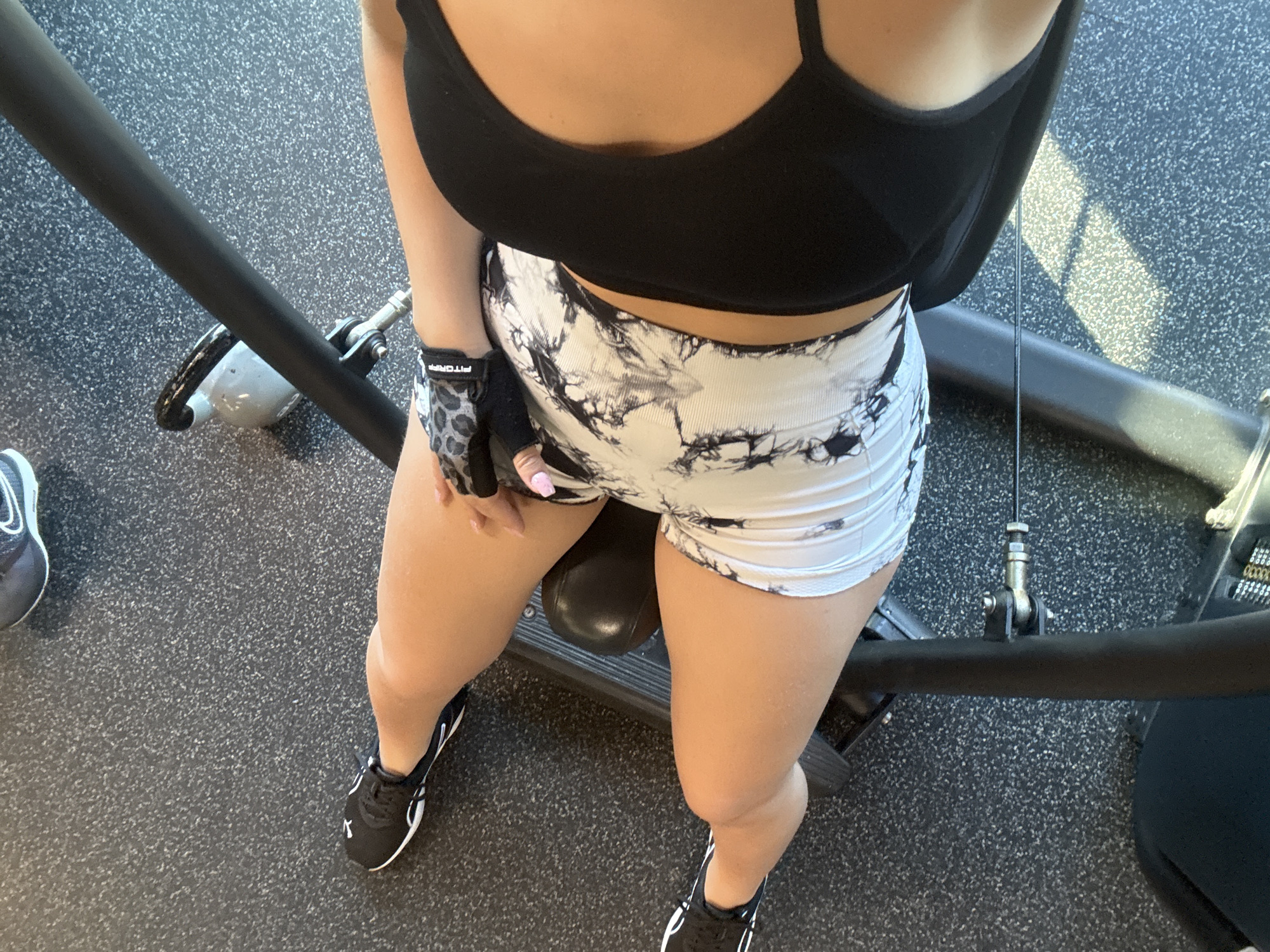 An overhead, high-angle, full-shot photograph captures a person, appearing to be female, from the thighs down, seated on a gym machine. The individual is wearing black athletic shoes with white accents, black and white tie-dye patterned shorts, and a black sports bra. A black and grey leopard print weightlifting glove is on the person's left hand, which is resting on their thigh. The background is a dark grey speckled gym floor, and a portion of a black metal gym apparatus is visible. A portion of a kettlebell is also visible to the upper right of the frame, on the gym floor. The scene is located in the City of Yonkers, United States of America.