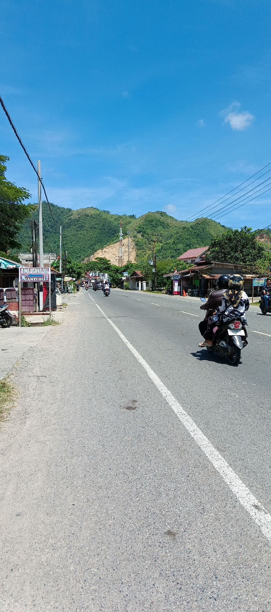 This is a photograph taken from a low angle, looking down a paved road in Rimajeunue, Indonesia. The road is asphalt with a white line dividing the lanes. On the left side of the road, there is a sidewalk made of gravel and concrete, with some grass growing on the edge. There are several small shops and stalls lining the road, including one with a sign that reads "BINA USAHA OIL GANTI OIL". A few motorcycles are parked on the side of the road.
In the middle of the road, there are several vehicles, including cars and motorcycles. One motorcycle is in the foreground, carrying two people wearing helmets. The background features lush green hills and mountains, with a clear blue sky above. There are also some power lines and a telecommunications tower visible on the hills. The overall scene depicts a typical day on a busy road in a rural Indonesian town, with a mix of commercial activity and natural landscape.