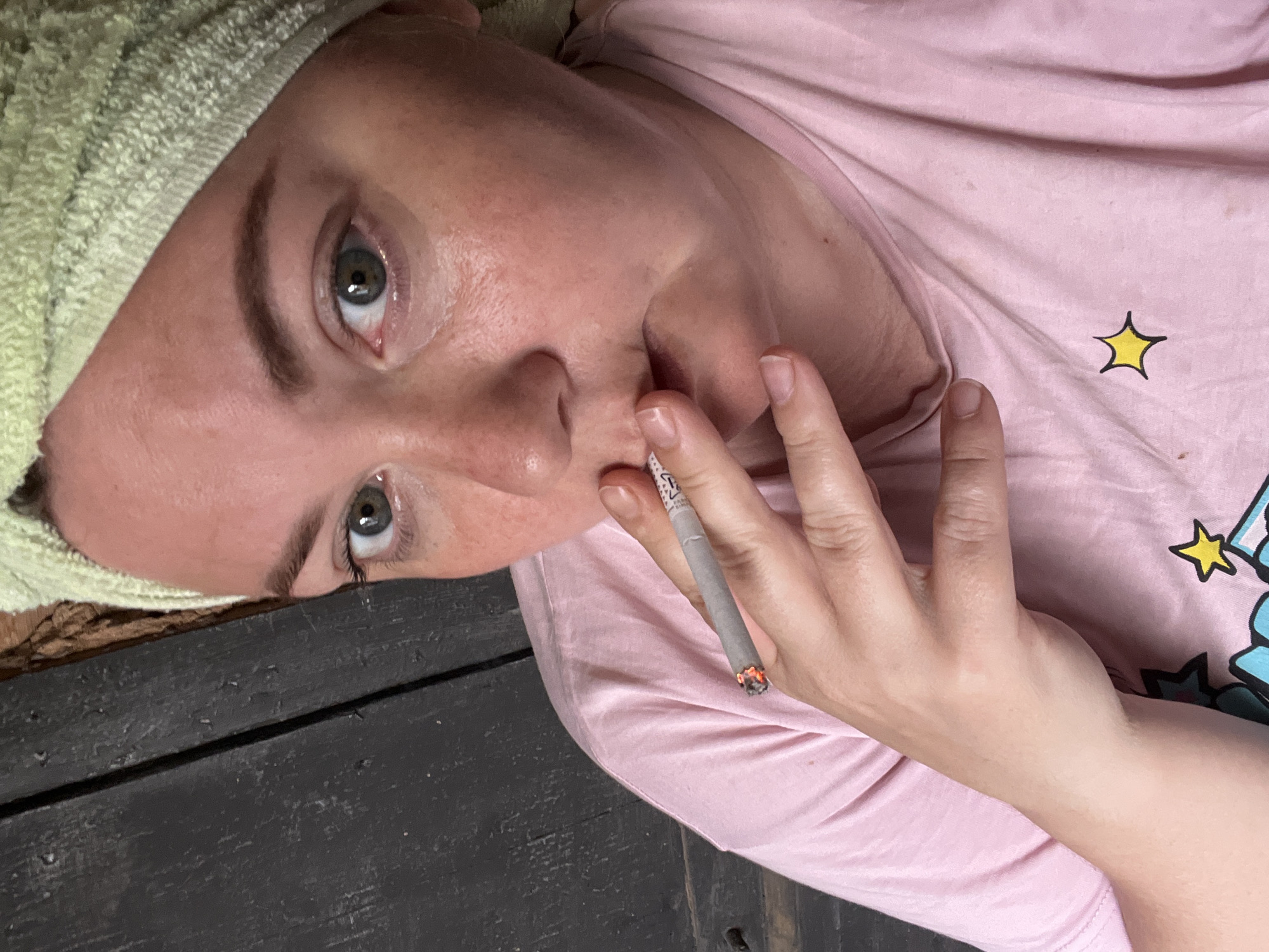 A woman with green eyes and brown eyebrows is seen from the chest up, holding a lit cigarette to her mouth. She is wearing a pink t-shirt with cartoon stars and a light green towel wrapped around her head. The background consists of dark wooden planks. The scene is indoors, possibly in a home in the city of Fokino, Russia. The lighting is natural, suggesting daytime.