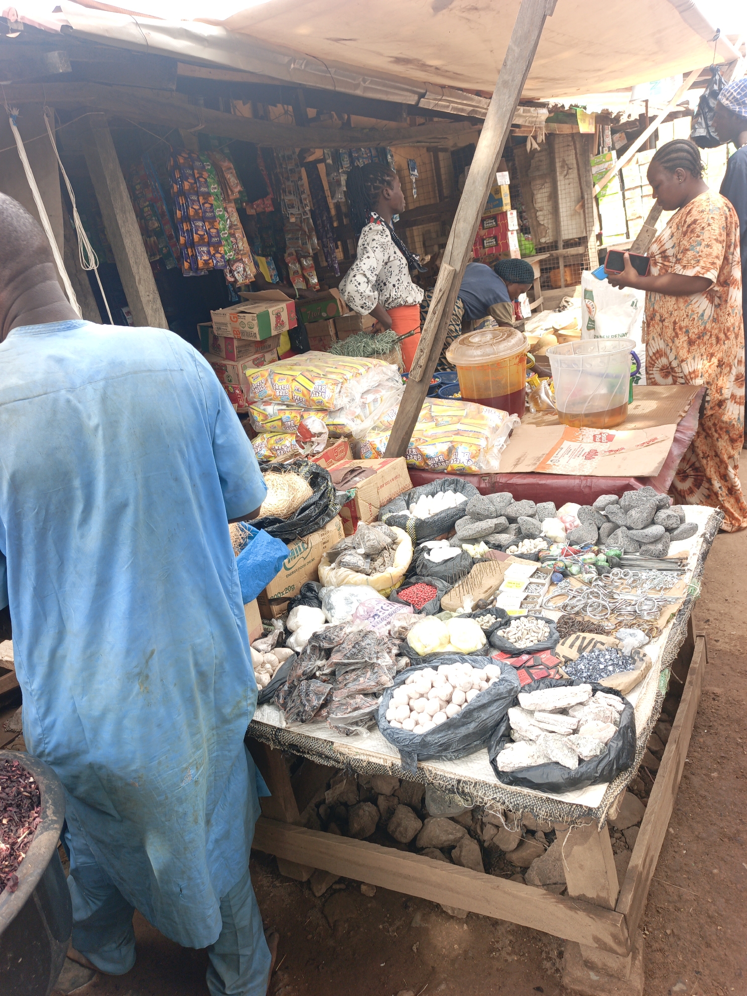 The image captures a bustling market scene in Akwanga, Nigeria. Under a makeshift shelter of wooden poles and a tarpaulin roof, a stall displays an array of goods.
A man in a light blue traditional outfit stands at the front of the stall. His back is to the camera. Behind him, various items are laid out on a table, including what appears to be foodstuffs, herbs, and other merchandise. A plastic container with a red liquid can be seen. Bags filled with goods are piled on the stall, and other packaged items are displayed on shelves behind the vendors.
Two women are examining the goods at the stall. The woman on the right has a phone in her hand. Several other people are visible in the background, some of whom appear to be customers.
The market stall is constructed of wood, with a makeshift base supported by rocks. The ground is dirt.