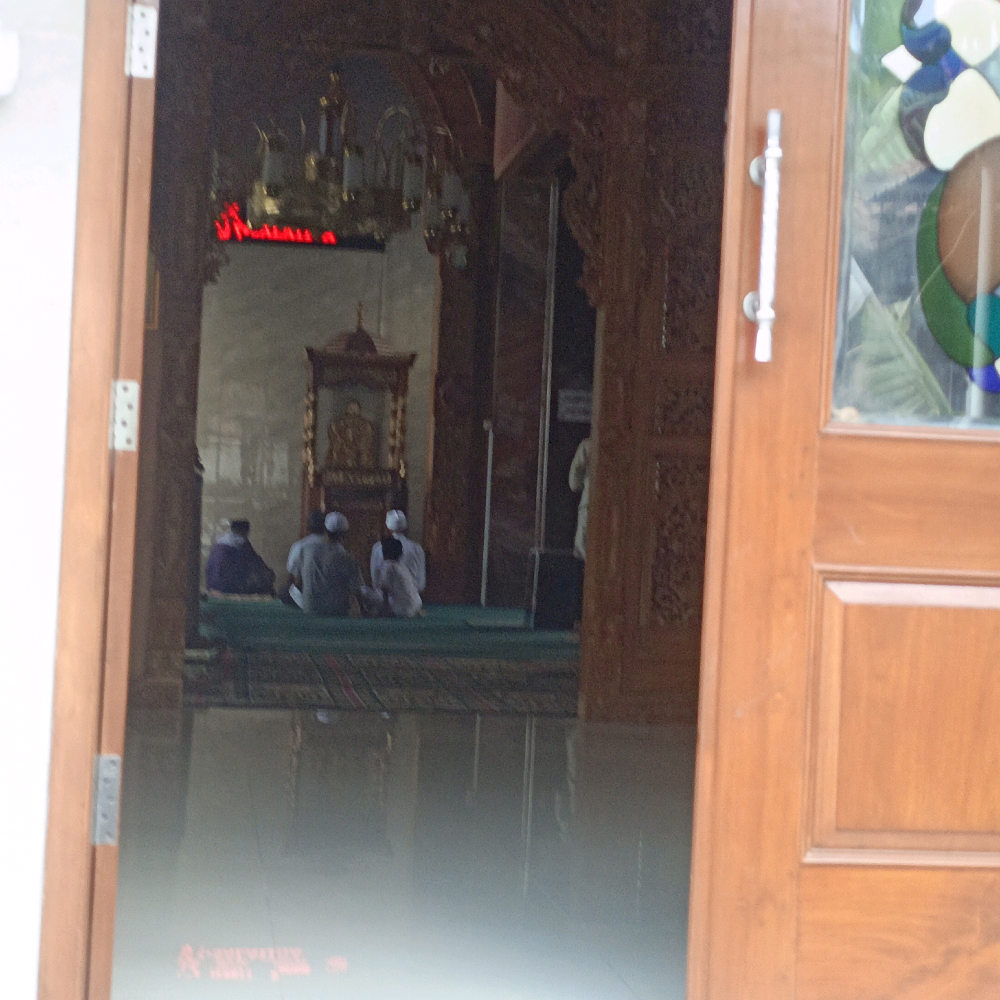 The image shows the entrance to a mosque in Salatiga, Indonesia. Through an open wooden door, we see the interior of the mosque where several men are sitting on green carpets in prayer. The men are wearing white caps and traditional attire. In the background, there is an ornate structure with carvings, possibly a minbar or a decorative element of the mosque. Red Arabic script is visible on a panel above the prayer area. The door itself is made of polished wood, with a silver handle. A stained-glass window with colorful geometric patterns is visible on the right side of the door. The overall impression is of a serene and devotional atmosphere within a place of worship.