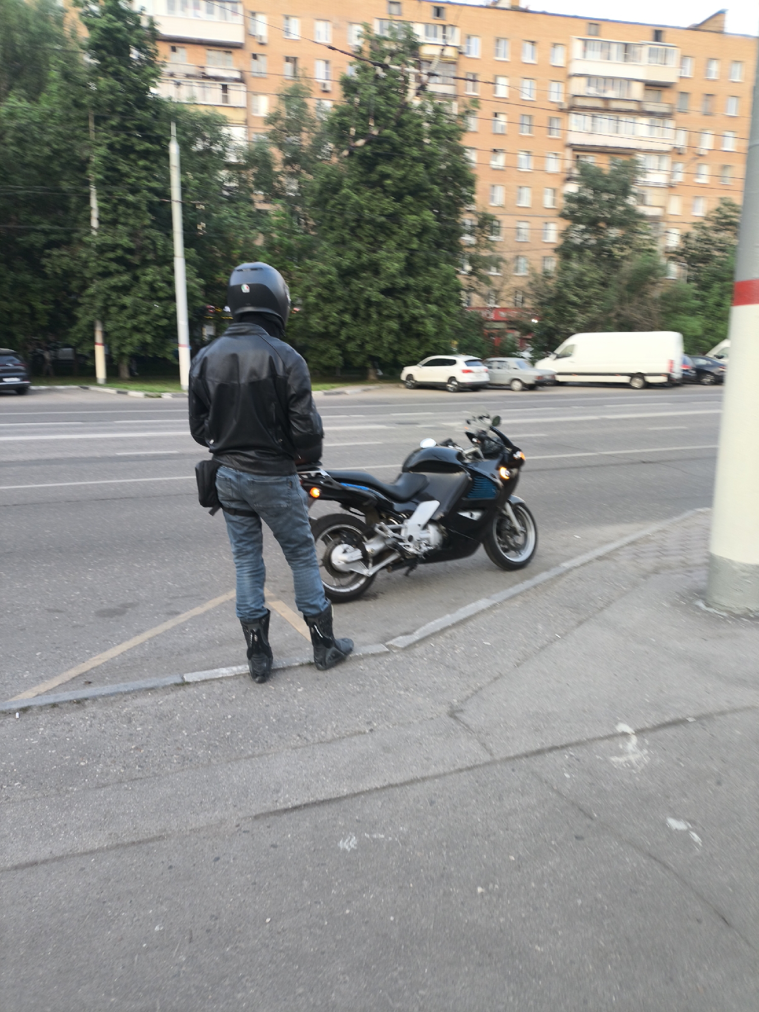 The photograph shows a man in a black helmet, a black leather jacket, blue jeans, and black boots standing on a sidewalk next to a black motorcycle. The man is facing away from the camera, looking toward a street with several vehicles, including a white van and a white car. Trees and a multi-story building are visible in the background. The location appears to be a city street. The city and its location are unknown.