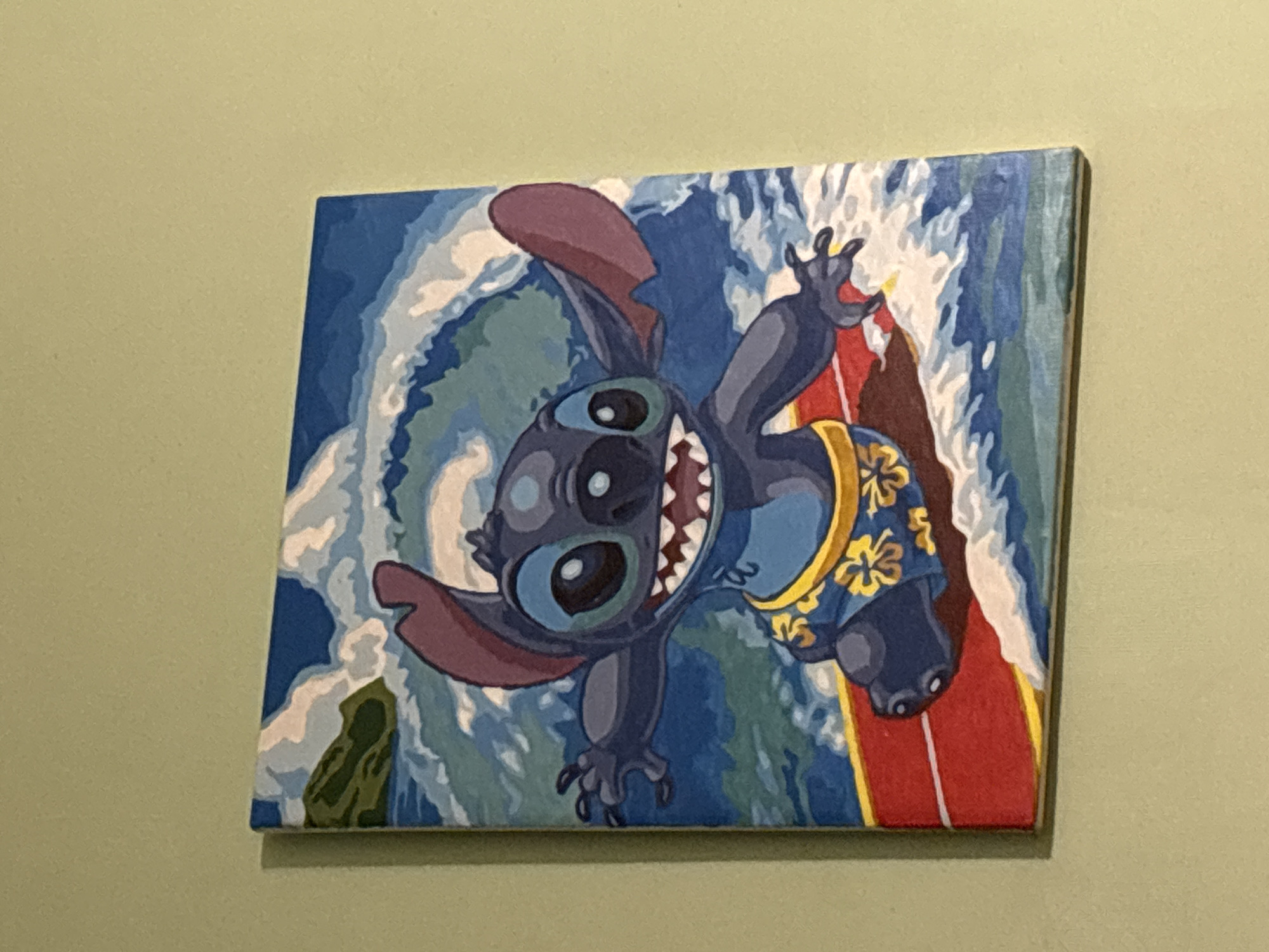 A framed painting hangs on a pale green wall. The painting depicts the Disney character Stitch surfing a wave. Stitch is dark blue with large eyes, prominent ears, and a wide smile revealing his teeth. He is wearing a blue shirt with yellow floral patterns. He is positioned on a red surfboard with yellow accents. The wave is stylized with blues, greens, and whites. The canvas wraps around a wooden frame. The painting appears to be indoors. There is no information about the location in the city of Vsevolozhsk, Russia.