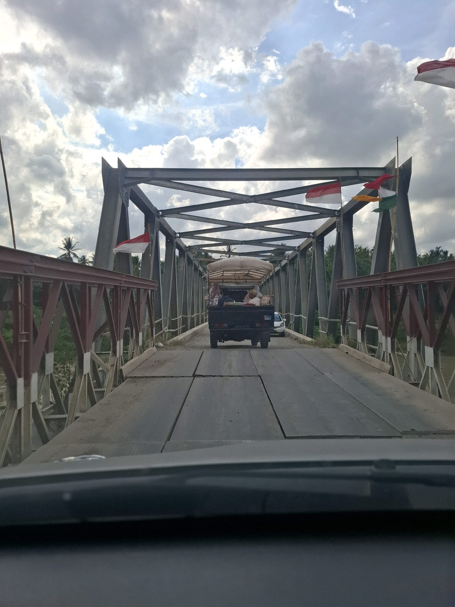 An eye-level, outdoor, medium shot captures a view from inside a vehicle looking forward at a bridge. The bridge, constructed of metal trusses, spans a body of water that is not fully visible. On the bridge's roadway, a dark-colored pickup truck is moving away from the camera, its bed filled with a large, tan-colored tarp-covered load. The truck has several people in its cab. A white car is visible behind the truck. Several Indonesian flags, red and white, are affixed to the bridge's structure and wave in the breeze. Palm trees and other dense foliage are visible on either side of the bridge, suggesting a tropical environment. The sky above is partly cloudy with patches of blue. The foreground of the image is occupied by the dashboard and windshield of the vehicle from which the photo is taken. The image depicts the bridge in Teupinreudeup, Indonesia.