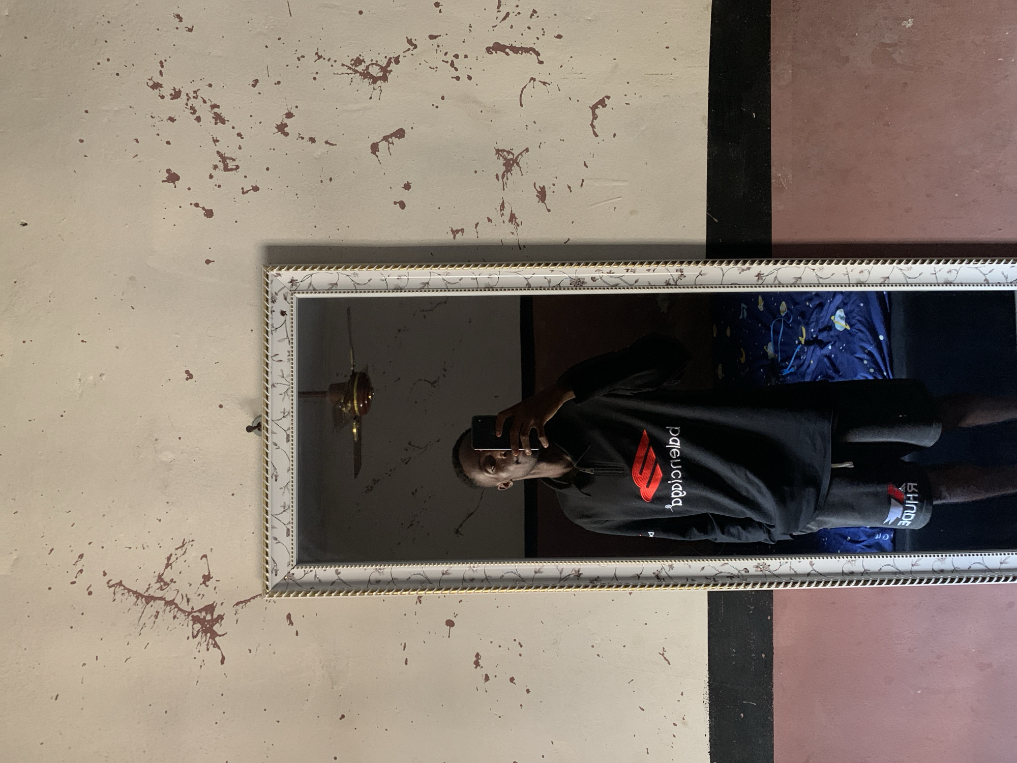 The image, originally rotated 90 degrees counter-clockwise, depicts a person taking a mirror selfie indoors. The individual, a male, is seen from the chest up, holding a smartphone to capture his reflection. He is wearing a black hoodie with a red and white logo that, when viewed in reverse in the mirror, appears to be "BPLGICNBD" (likely "BALENCIAGA" or a similar brand). He also wears black shorts with the reversed text "RHUDE" clearly visible on them.
The mirror features a decorative white frame with a golden inner border and an intricate floral or vine pattern. Reflected within the mirror, above the person, is a dark-colored ceiling fan, and below him, a blue fabric with white patterns, possibly bedding.
The wall surrounding the mirror is a light cream or beige color, extensively marked with numerous irregular brown or reddish-brown splatters and streaks. To the right of the mirror, the wall transitions into a distinct vertical black stripe, followed by a solid reddish-brown section. The indoor setting, possibly in Lokoja, Nigeria, presents a somewhat distressed or lived-in aesthetic, with ambient, somewhat dim lighting.