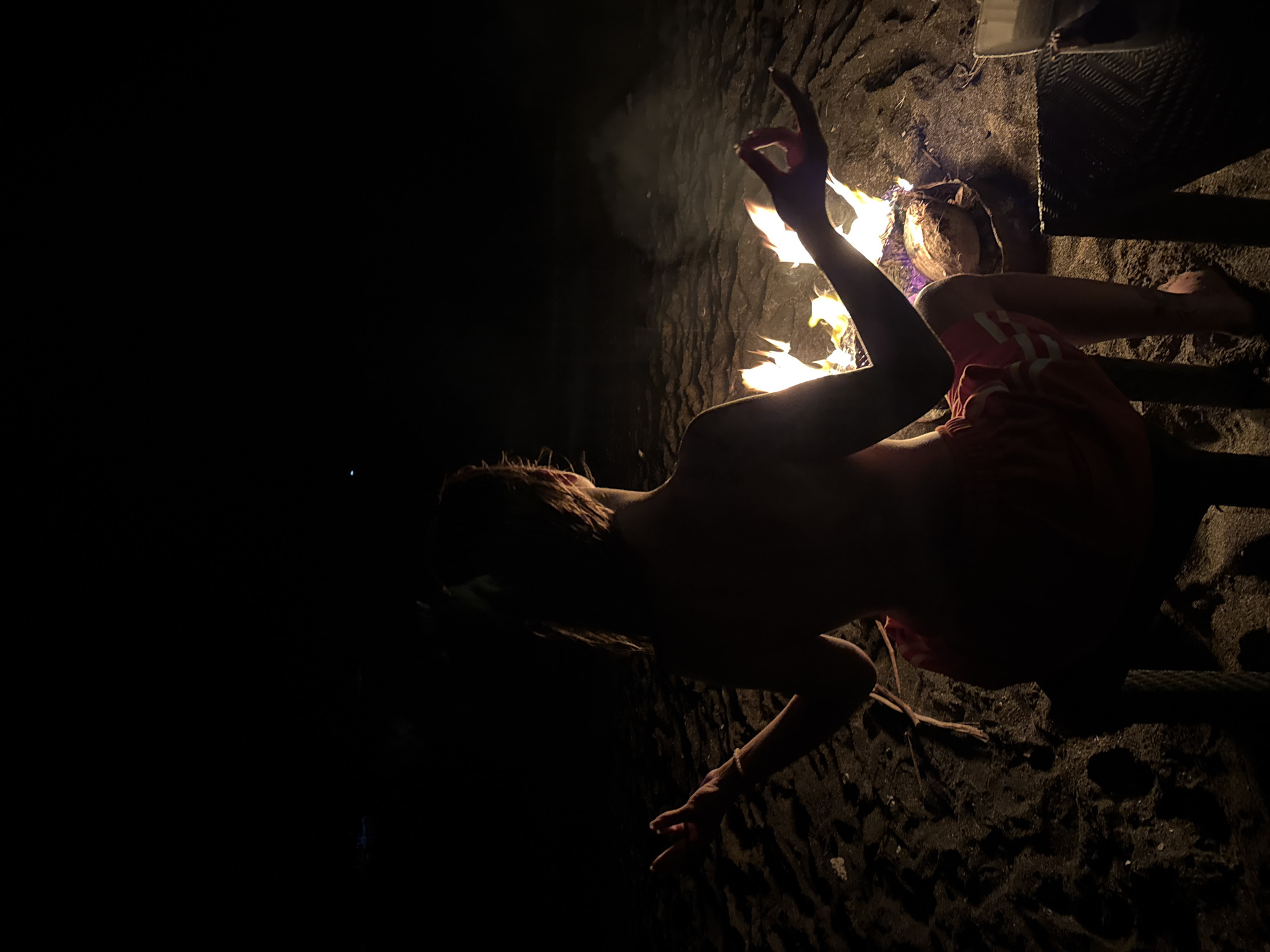 A person wearing red shorts and no shirt is seated on a black, sandy beach next to a fire at night in Unknown, Indonesia. The person's right arm is raised, with fingers curled in a gesture. The area to the left of the person is shrouded in darkness.