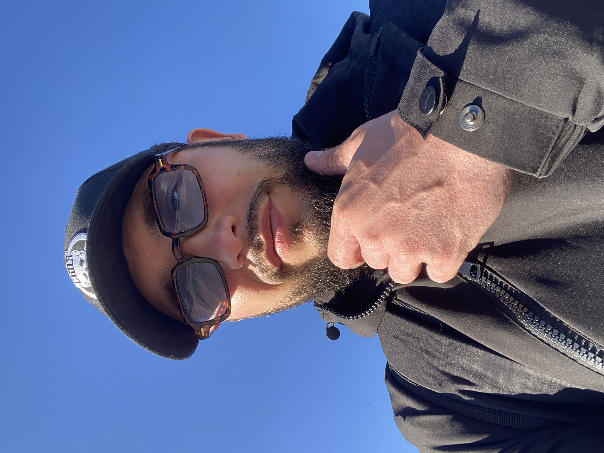 A man is taking a selfie outdoors under a bright blue sky. He is wearing a black baseball cap with a white logo, tortoise-shell framed glasses, and a black jacket. His right hand is touching his chin and beard. The image shows a close-up of his face and upper torso. There is no indication of the city of Koskolovo, Russia in the image.