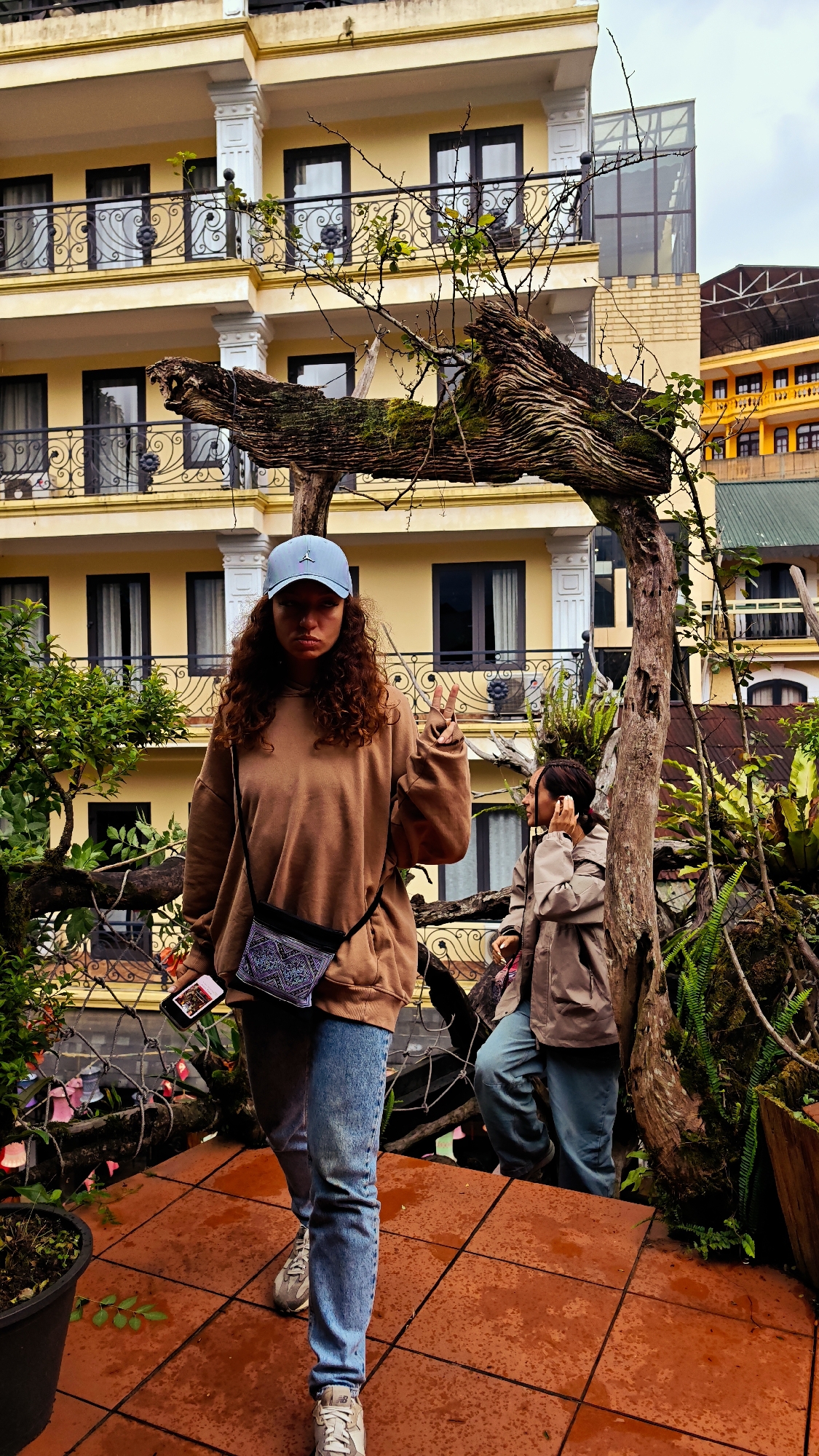 A full shot photograph captures a woman facing forward in Sa Pa Ward, Vietnam. She wears a light blue baseball cap, brown hoodie, blue jeans, and gray sneakers. A patterned cross-body bag and a phone in her left hand accessorize her outfit. Her right hand is raised, forming a peace sign. Behind her is another woman on stairs, wearing a tan jacket and blue pants.
The background is a multi-story yellow building with black metal railings on the balconies. A weathered wooden archway is partially in front of the building. The foreground and setting include plant life and a reddish-brown tiled surface.