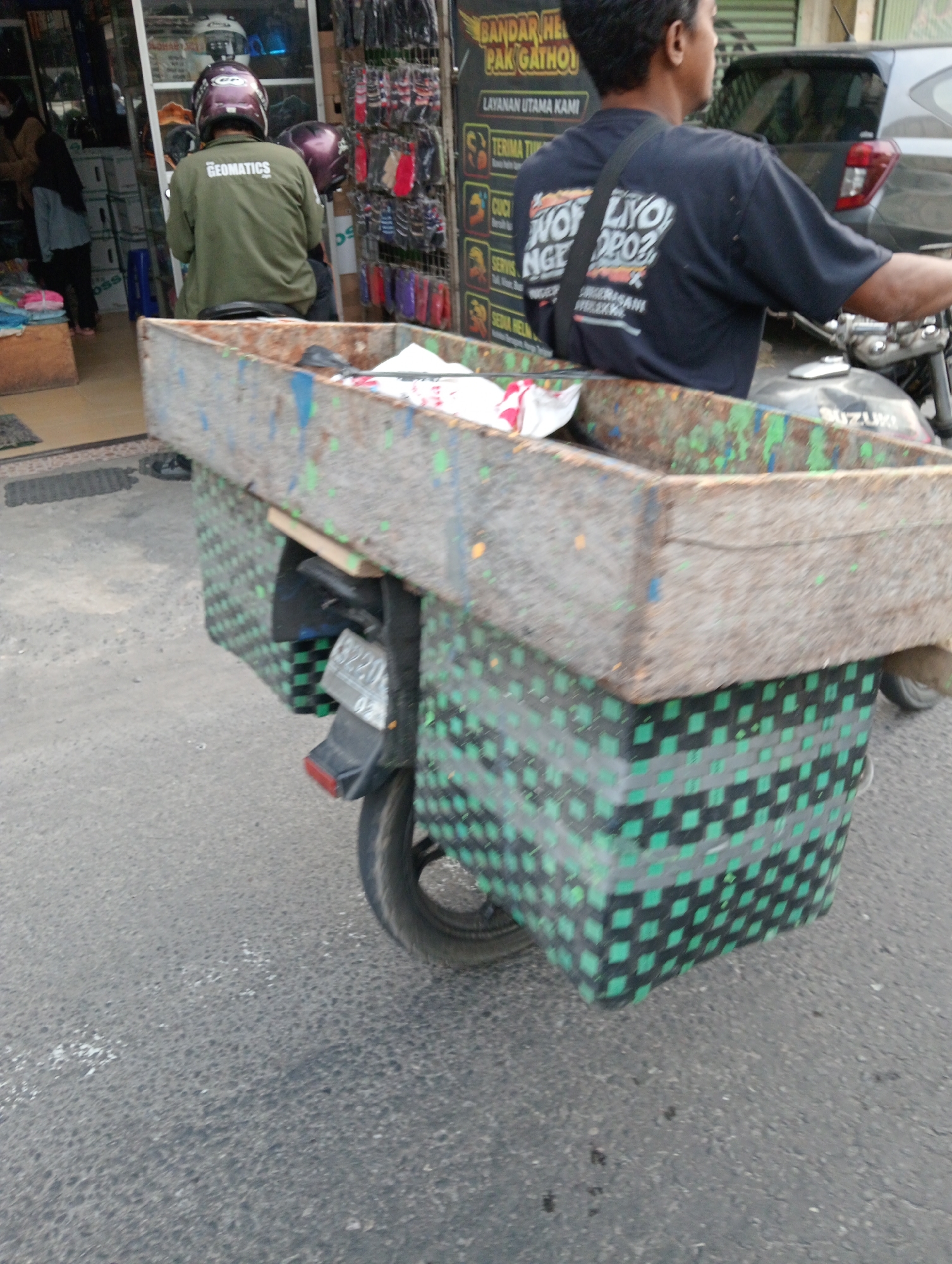 The image shows a two-wheeled vehicle, likely a motorcycle, customized with a large wooden box attached to the rear. The box is open at the top and appears to be carrying some items, possibly for delivery or transport. The box has a distressed, weathered look, with visible paint splatters in various colors, including blue, green, and yellow. The sides of the box are reinforced with a woven material, predominantly in black and green checkered patterns. A license plate, partially visible, is mounted on the rear of the motorcycle, below the box.

A person is seated on the motorcycle, facing away from the camera. They are wearing a dark-colored t-shirt with text and graphics on the back, and a black strap is visible across their chest. A maroon helmet is worn by the rider. In the background, to the left, a person wearing a light-colored jacket and a black hijab is visible, partially obscured by the motorcycle. Further in the background, a shop or storefront can be seen, with shelves displaying various items, including helmets and other accessories. A sign with text is mounted on the shop. The ground is paved with asphalt.

The scene appears to be on a street in Gulon, Indonesia, likely in a commercial or market area, given the presence of the shop and the general urban setting. The lighting suggests it is daytime.