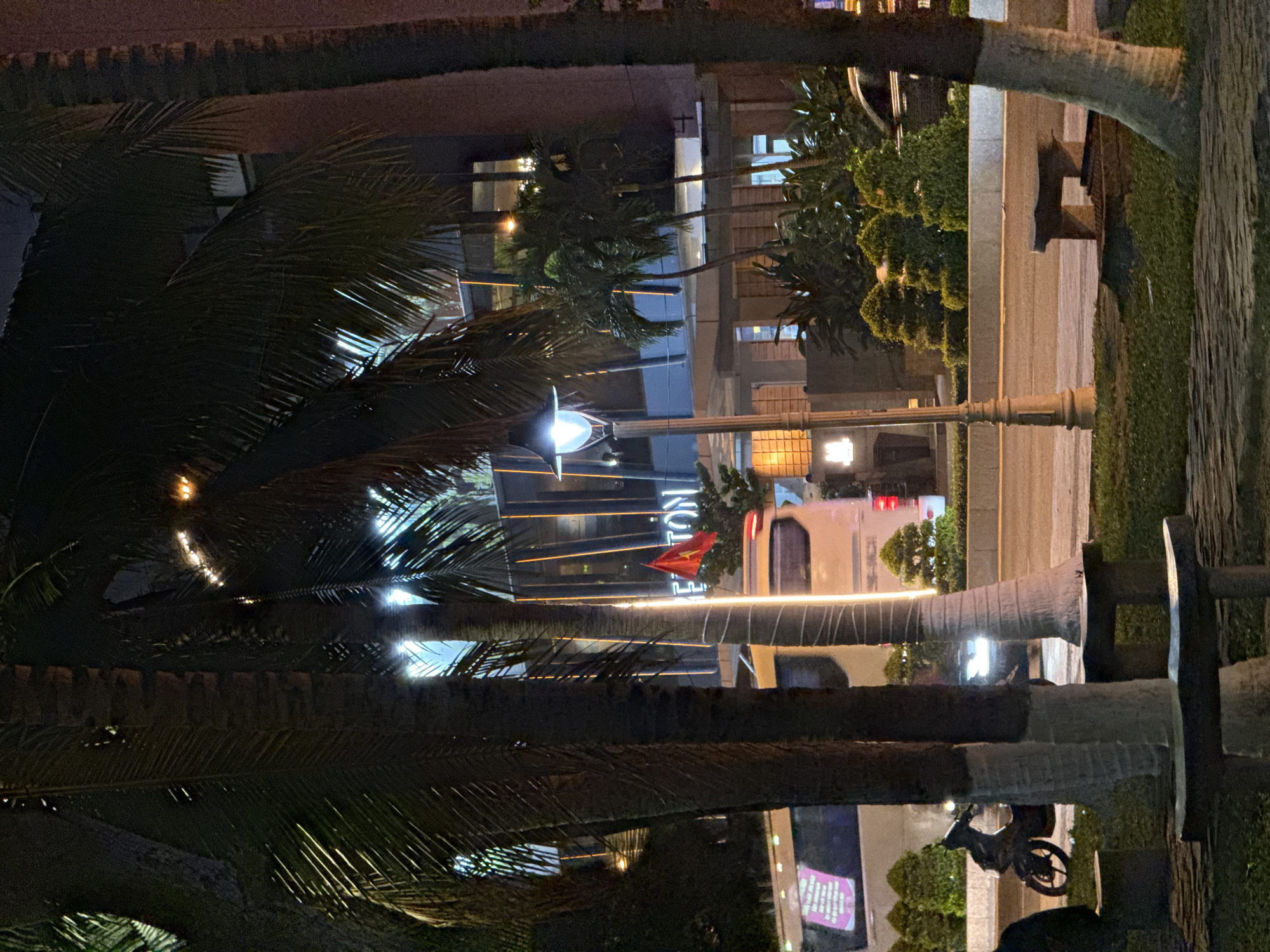 In Nha Trang, Vietnam, at night, a street scene unfolds with palm trees in the foreground framing a building. The building has a modern facade with vertical lights and large windows, suggesting a hotel or commercial establishment. A red flag, likely the Vietnamese flag, is visible attached to the building. In front of the building, a white van with text on its side is parked, and a scooter is on the road. A lamppost with an illuminated lamp stands near the sidewalk. The ground is paved with stone and grass. The overall impression is an urban night view with a blend of natural and architectural elements.