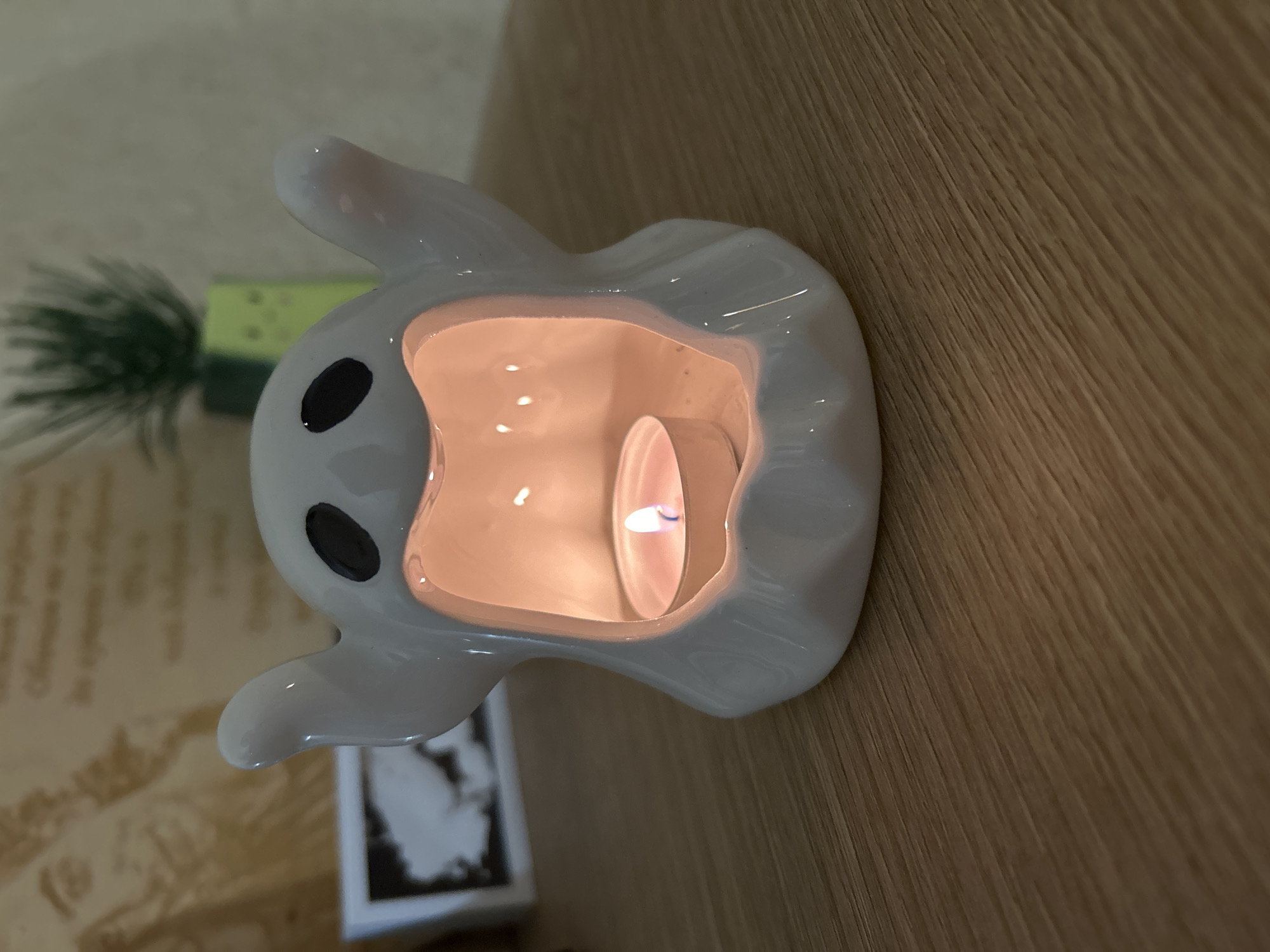 A white, ghost-shaped ceramic tea light holder, reminiscent of the character Zero, is prominently displayed in this indoor scene. The holder features large black eyes and a friendly, cartoonish design, with a lit tea light candle nestled inside. The candle emits a warm, orange glow, illuminating the interior of the ceramic ghost and casting a soft light on its surface.
The holder rests on a polished wooden surface, which exhibits a clear grain pattern, suggesting a table or shelf. To the left and slightly behind the ghost, a blurry artificial green plant, possibly a small pine or fir, is visible, adding a subtle touch of greenery to the background. Further back and to the left, another blurry object with a light-colored, textured surface, possibly a book or decorative item with faint patterns, can be discerned.
The overall lighting is dim, emphasizing the bright, flickering flame of the tea light candle within the ghost holder. The image is rotated 90 degrees counter-clockwise, orienting the wooden surface vertically. There is no discernible text in the image. The scene conveys a cozy and possibly festive or comforting atmosphere, likely in a home setting in Sofia, Bulgaria.