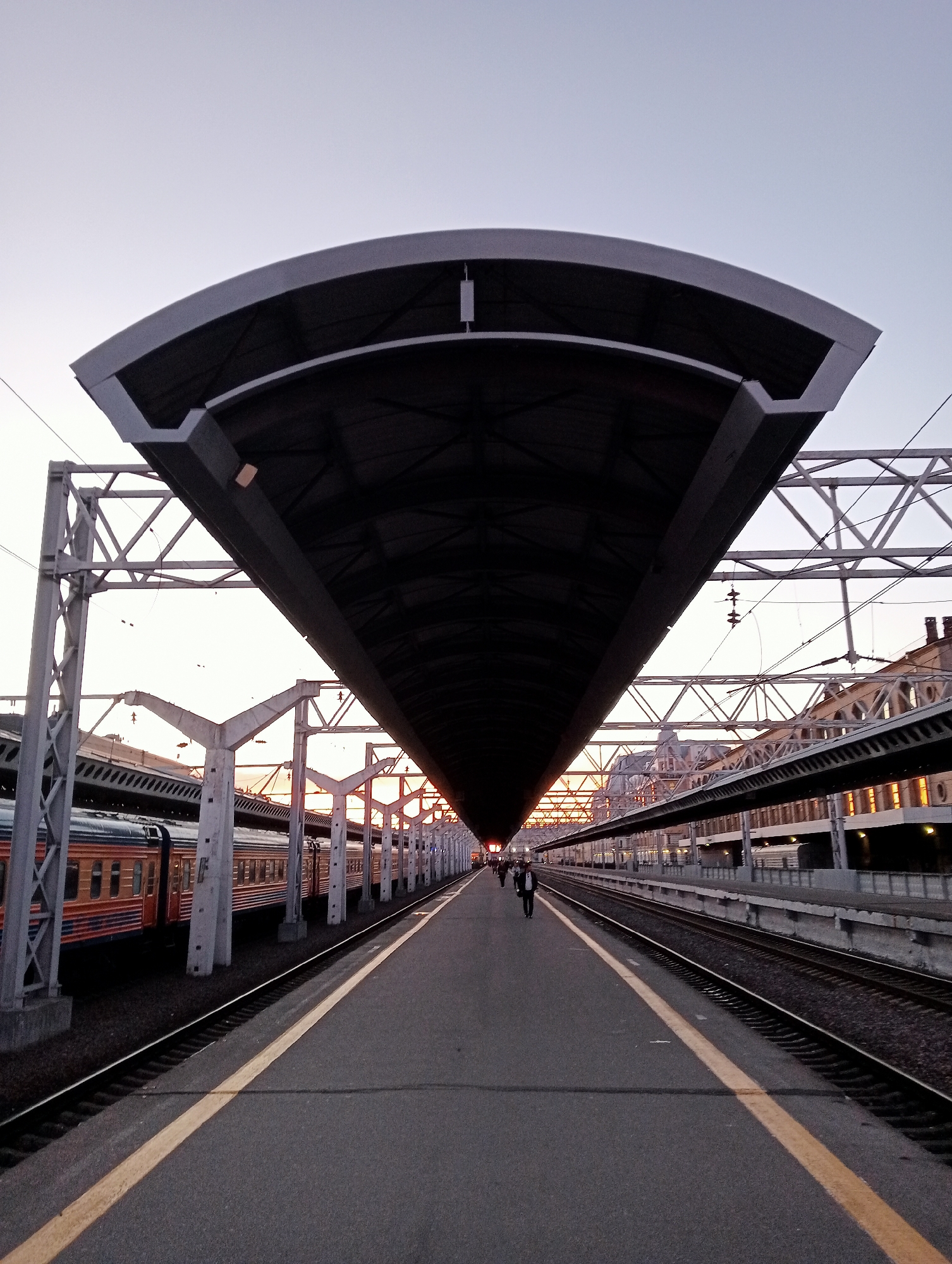 The image captures a train station platform at twilight, likely in Saint Petersburg, Russia. A long, arched roof structure dominates the foreground, casting a shadow over the empty platform. To the left, a train with orange and blue carriages is parked along the tracks. Several figures are walking on the platform in the distance, their forms silhouetted against the dim light. The sky is a pale gradient of grey and white, suggesting the early stages of evening or late dawn. The platform itself is asphalt with a distinct yellow line marking the edge of the tracks. Industrial-looking metal supports and overhead electrical lines are visible, typical of a functioning railway station. The architecture of the buildings lining the opposite platform, with their arched windows, hints at historical significance.