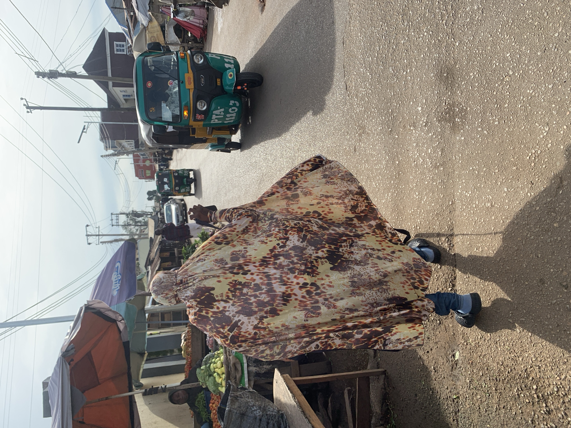 The image depicts a street market scene in Jos, Nigeria. In the foreground, a person walks away from the camera, wearing a flowing, patterned garment and blue jeans. Their feet are clad in black slip-on shoes. The person is casting a long shadow on the asphalt road, which is littered with small stones and pebbles. To the left of the person, market stalls are visible, with an orange umbrella providing shade over produce, including tomatoes and leafy green vegetables. A vendor is seated behind the produce. Further down the street, a green and yellow auto-rickshaw, identified by the license plate "PTA 11103," drives past, its shadow also stretching across the road. Another auto-rickshaw is seen further in the distance. The background shows buildings and overhead power lines, suggesting an urban or semi-urban environment. The sky is overcast with bright sunlight.