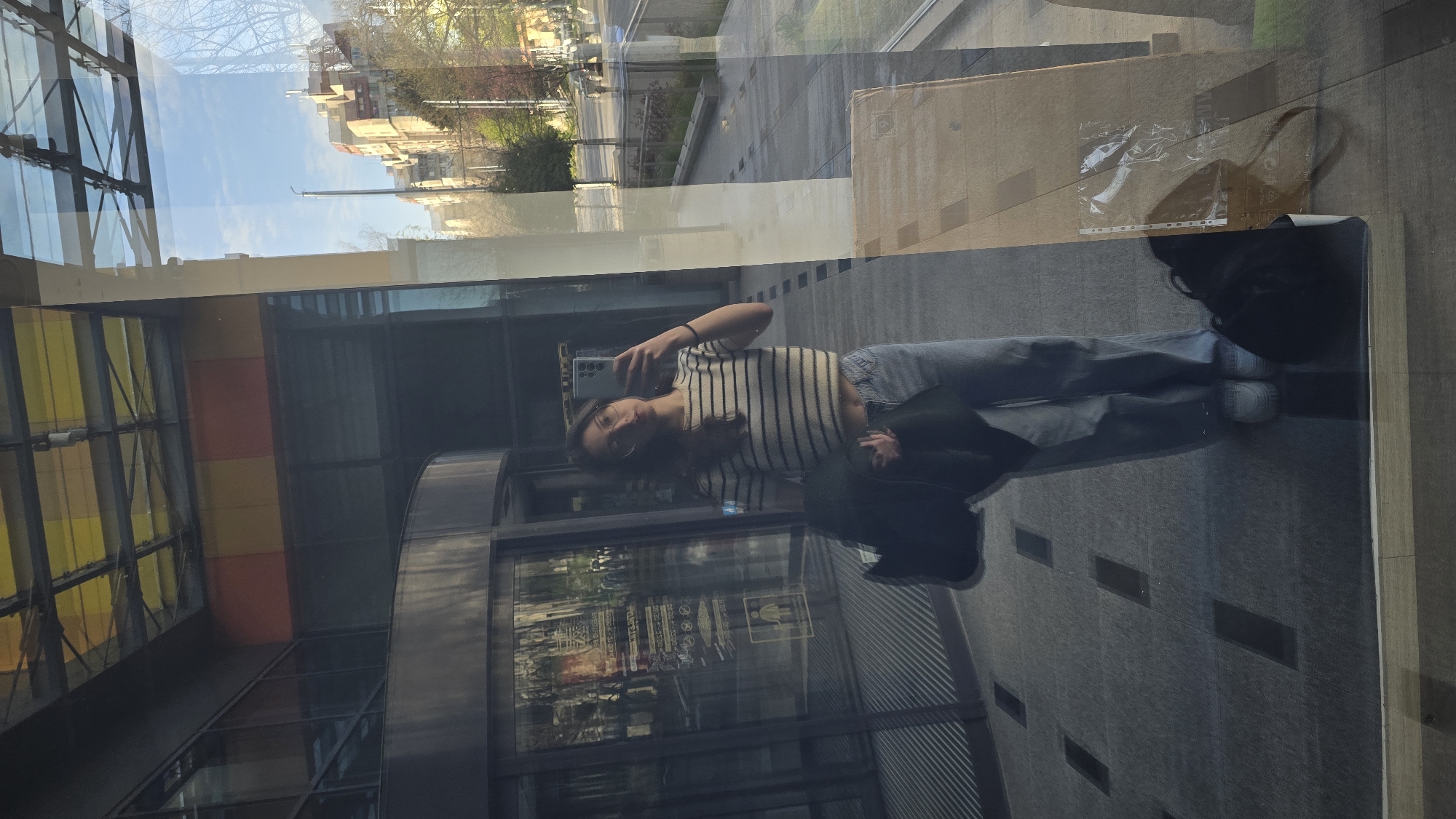 The image shows a reflection of a person taking a photo of themselves in front of a glass facade. The facade reflects a building and a street scene, potentially in Stara Zagora, Bulgaria. The person is wearing a striped top and jeans. A jacket or bag is held in one hand. In the reflection, a structure with yellow, red, and transparent panels can be seen. The overall setting appears to be an urban environment.
