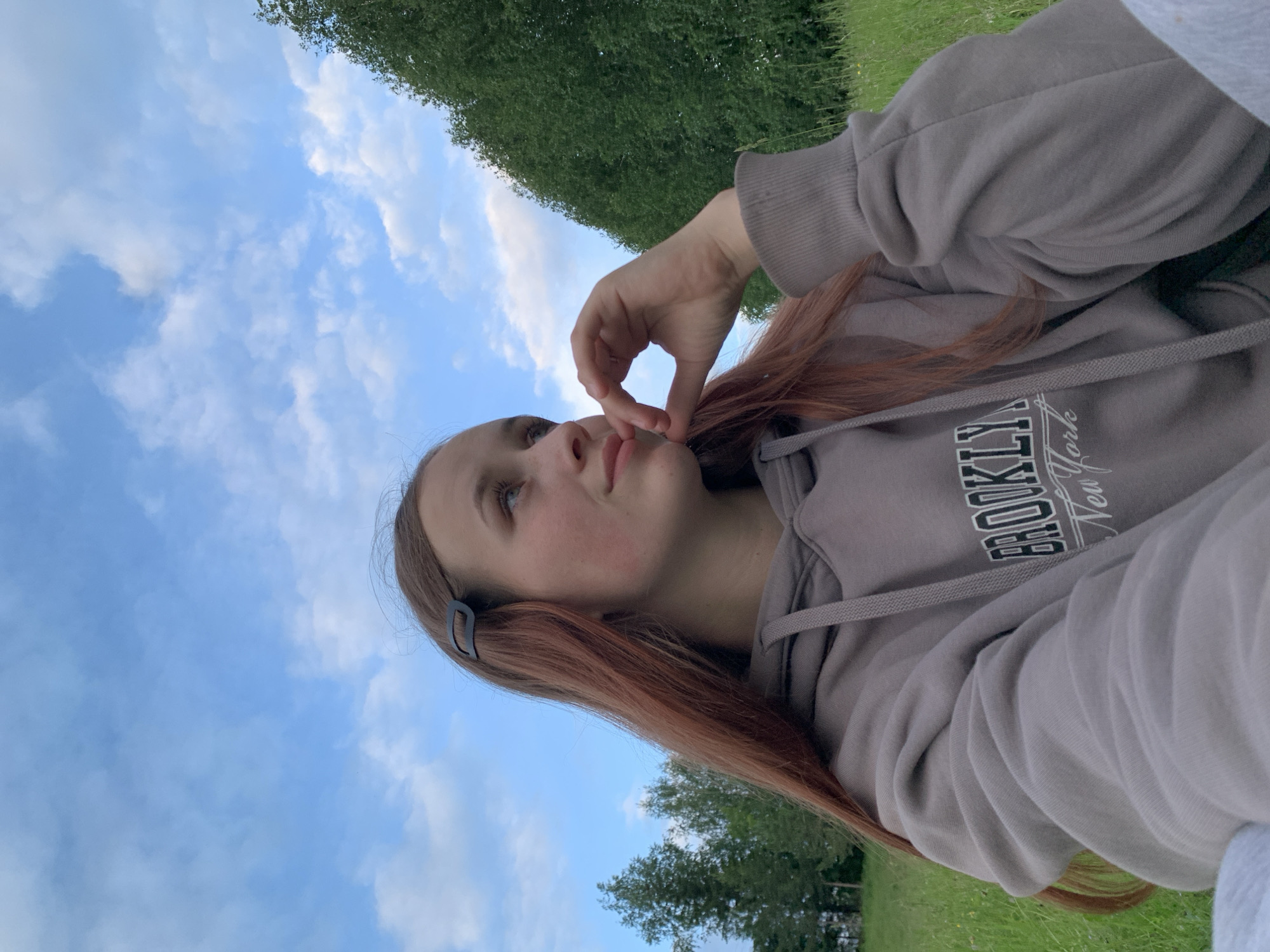 An individual, likely female, with long reddish-brown hair secured by a black hair clip on the right side of her head, is depicted in an outdoor setting in Фоки, Russia. She wears a grey hooded sweatshirt featuring the text "BROOKLYN New York" on the front. Her head is turned to her right, and her gaze is directed upwards and to the right. Her right hand is positioned with fingers touching her chin. The subject is situated in a grassy area, with dense green trees visible in the background on both the left and right sides of the frame. The sky is blue with scattered white cumulus clouds. The scene is illuminated by natural daylight.