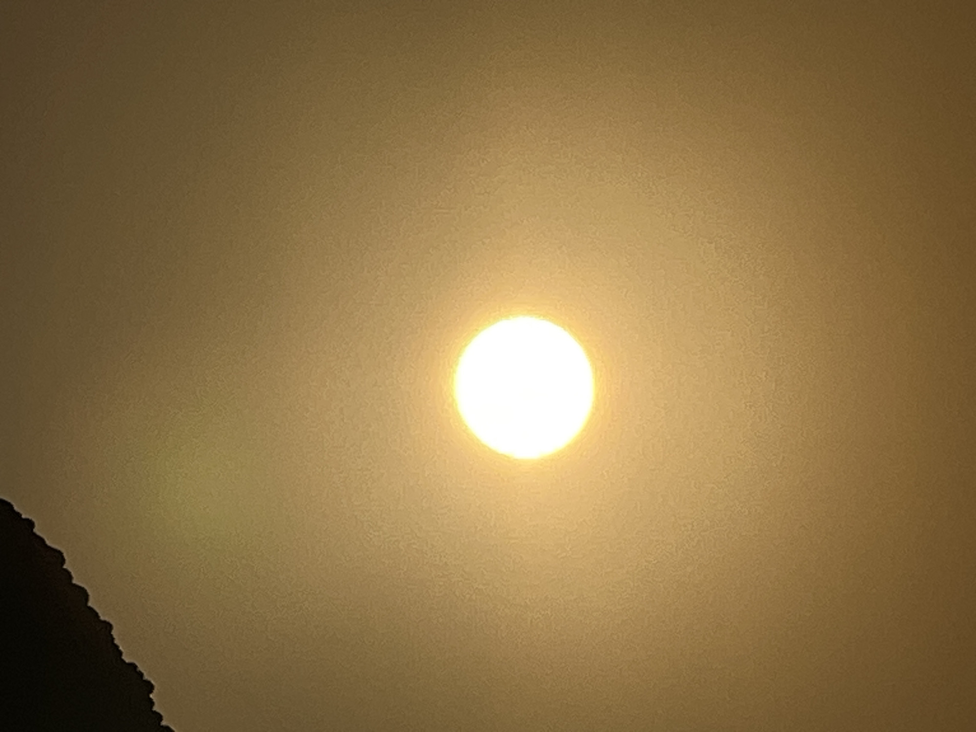 A bright, slightly blurred circle, presumably the sun, is centered in a hazy, golden-yellow sky. The sky occupies most of the frame, with a dark, jagged edge visible in the upper left corner, likely representing a shadow or an object obscuring the sky. The overall lighting suggests either dawn or dusk, or a day with significant atmospheric haze. There are no people or specific objects identifiable within the scene. The location in Kuta, Indonesia, is not visually evident from the image itself; this information is external context.