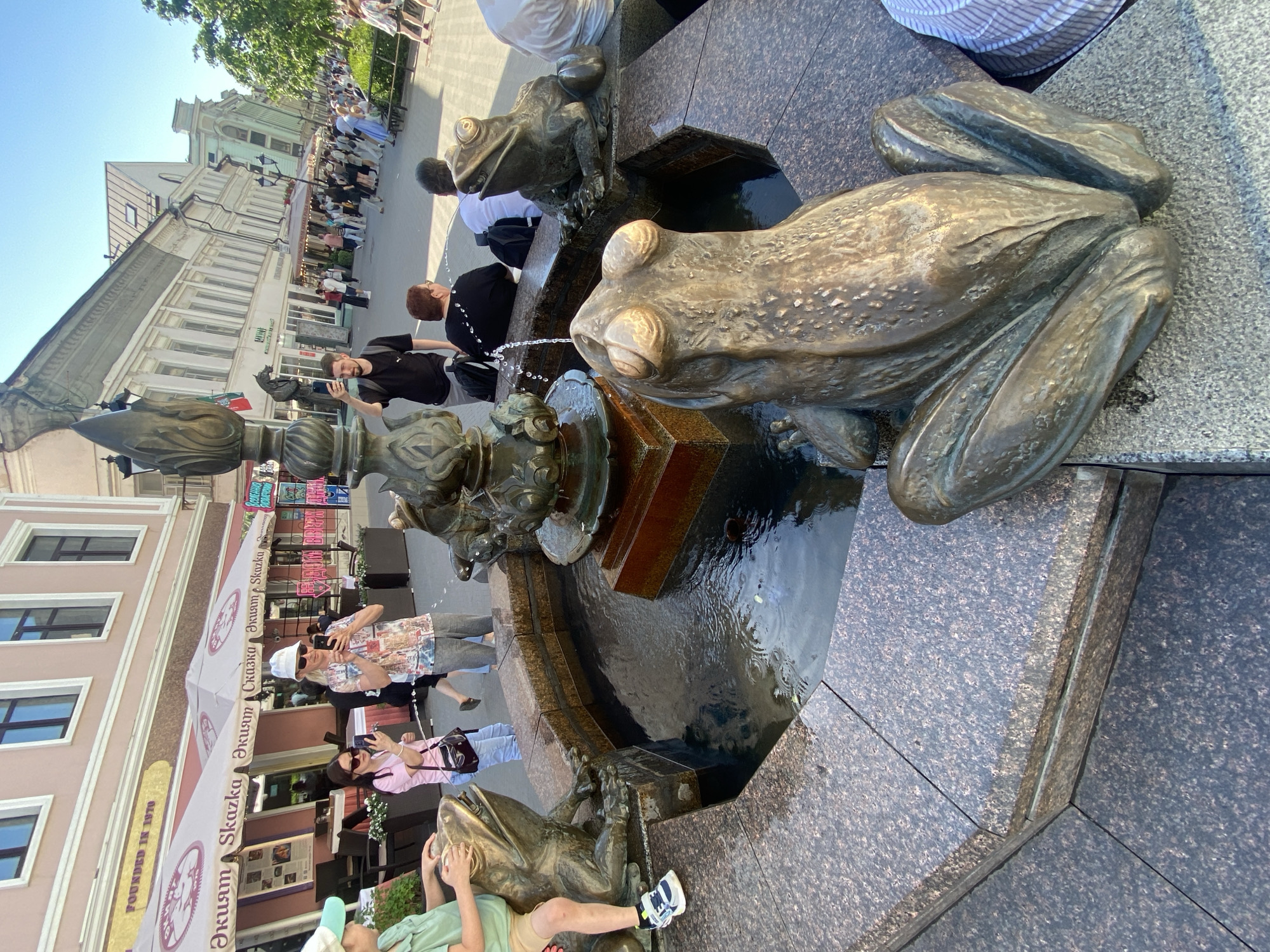This outdoor image depicts a frog fountain in the city of Kazan, Russia. The fountain is constructed of dark gray granite and features four bronze frogs. One frog is positioned prominently in the foreground, appearing large and detailed. Two smaller frogs are placed around the fountain's central water spout, while the fourth frog is partially obscured in the bottom left corner of the image.
Several people are present, mostly engaged in taking photos of the fountain. Some are standing nearby, while others are slightly further away, along what appears to be a pedestrian street. Behind the fountain, a restaurant with an awning reading "Skazka" is visible. Buildings line the street, and a group of people can be seen walking in the distance. The sky is blue, suggesting a sunny day. A child is sitting near the fountain with their feet dangling.