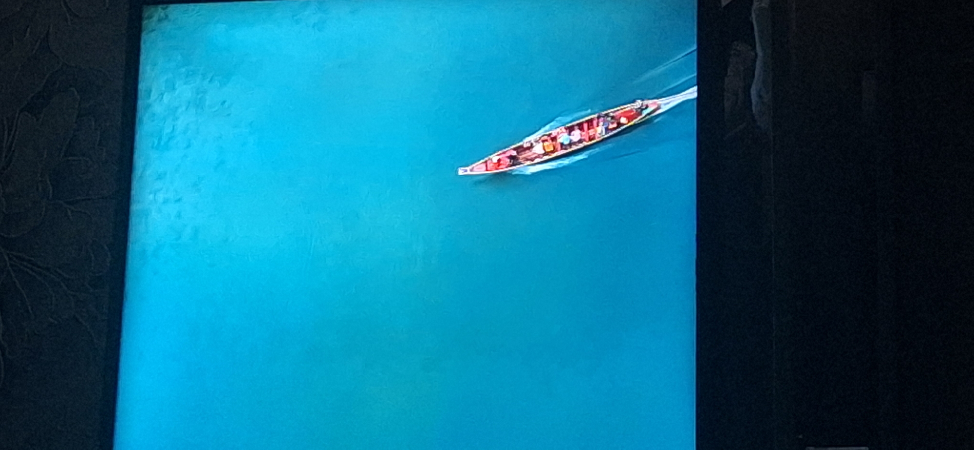 An aerial view captures a long, narrow red canoe with multiple occupants navigating through a vast expanse of bright blue water. The canoe appears to be in motion, indicated by the wake trailing behind it. The water is calm and smooth, reflecting the light and creating a serene atmosphere. The top edge of the image shows a dark, textured surface, possibly a ceiling or wall, with a subtle floral pattern. The bottom portion of the image is dominated by a dark, solid area, likely the edge of a screen or display. No specific landmarks or recognizable structures of Bila Tserkva, Ukraine are visible in this image.