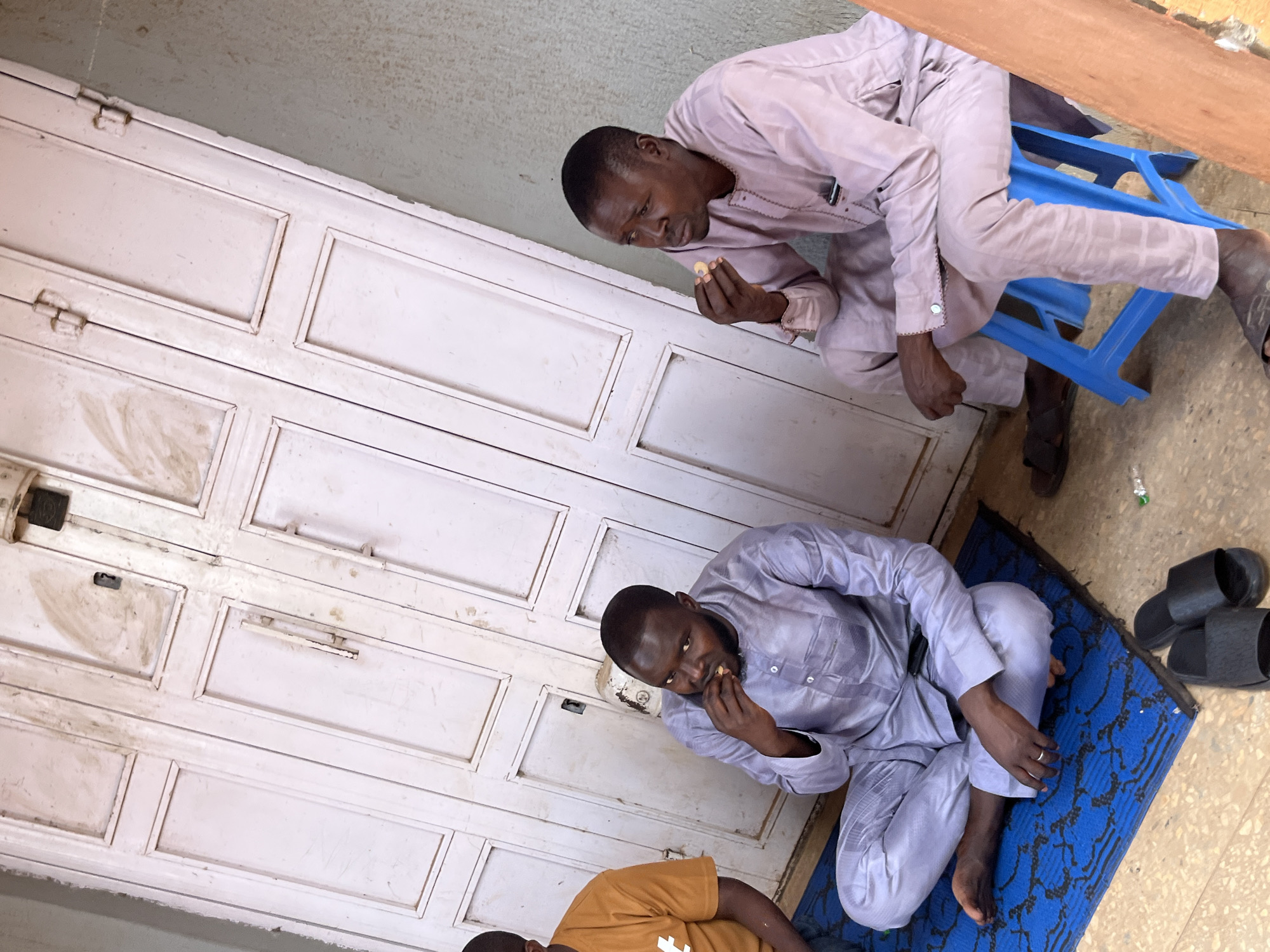 This tilted, eye-level shot captures three men in a casual indoor or sheltered outdoor setting, likely in Jos, Nigeria, during daylight hours.
In the midground, a man with a beard sits cross-legged on a blue mat adorned with a dark pattern. He wears a light grey traditional tunic and matching trousers, and a ring is visible on his left hand. He is bringing a small, light-colored food item to his mouth. Next to the mat, a pair of black slide sandals rests on a concrete-like floor. Partially visible in the lower foreground is another man in a mustard yellow t-shirt, also on the blue mat, facing away from the camera.
In the upper right, a third man sits on a bright blue plastic stool. He wears a light purple tunic and matching pants, and is also shown bringing a small, light-colored food item to his mouth. His feet are clad in dark sandals. Above him, a wooden plank or surface is partially visible, suggesting a table or architectural element.
The background is dominated by a white, multi-paneled door that appears worn, dirty, and features visible hinges and latches. The overall impression is one of a relaxed moment, possibly a break or informal gathering, as the men consume small snacks. There is no visible text on any of the subjects or objects.
