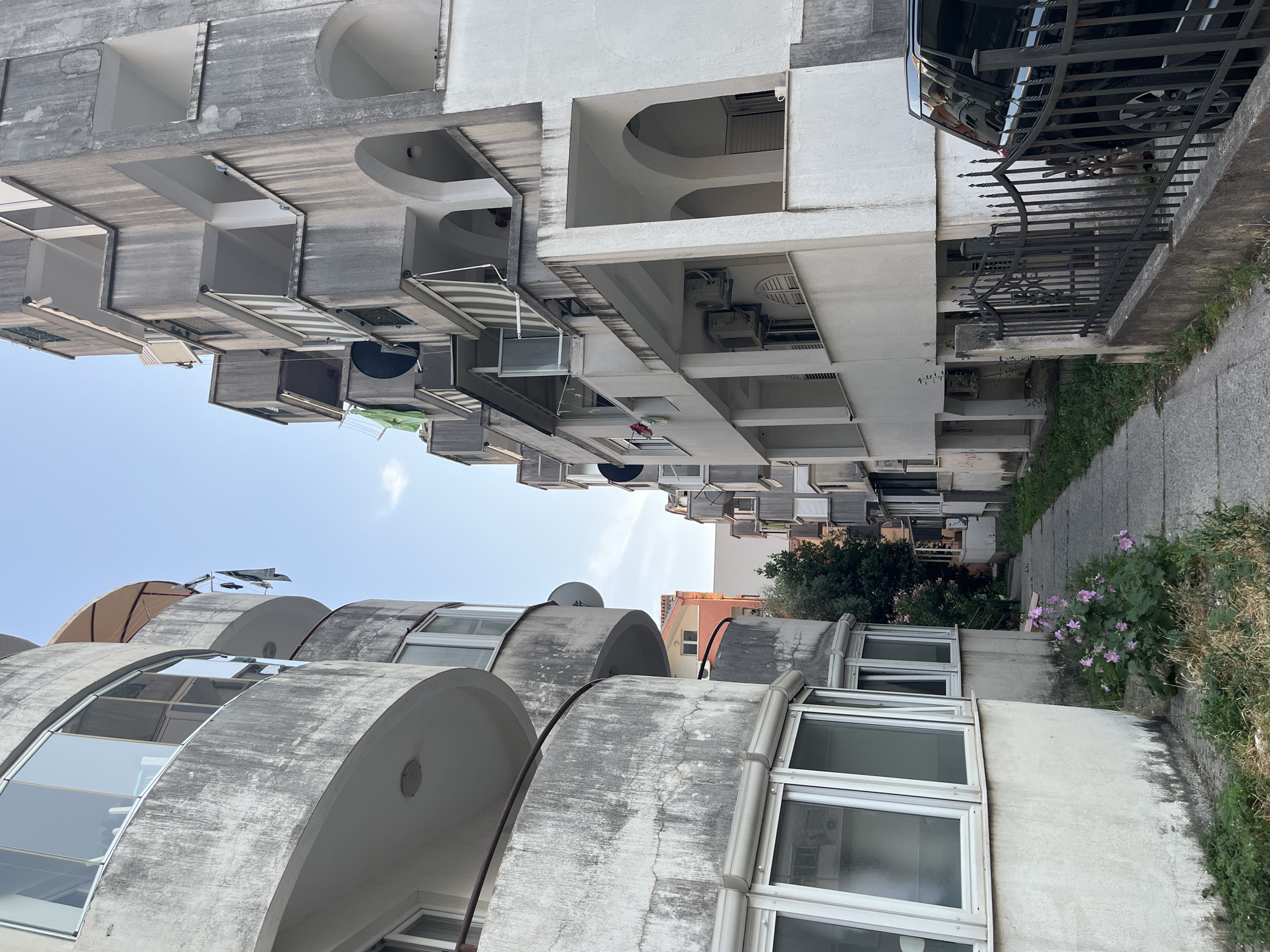 The image depicts a residential building with a unique architectural style, characterized by curved concrete balconies and protruding window frames. The building appears to be multi-story, with a mixture of white and gray concrete surfaces. Several balconies have awnings, some striped and others solid, suggesting sun protection. Air conditioning units are visible on some of the balconies.
The foreground features a set of curved balconies with large windows, some reflecting the sky. Behind these, the building rises further, showing more rectangular balconies with visible satellite dishes and what appears to be laundry hanging from a line on one of them. Further in the background, other buildings of varying architectural styles can be seen, including one with a pink facade.
To the right of the building, a narrow concrete pathway runs alongside it. Lush green vegetation and pink flowering plants grow along the edge of the pathway and the building's base, indicating a pleasant, possibly warm, climate. A black metal fence, ornate in design, separates a portion of the building from the pathway. A black vehicle is partially visible behind the fence, parked next to the building.
The sky is a clear, pale blue, suggesting it is daytime, likely late morning or early afternoon given the brightness. The overall scene conveys a sense of urban living in a Mediterranean or similar climate, with a blend of modern and perhaps older architectural elements. No people are visible in the image. There is no visible text in the image.